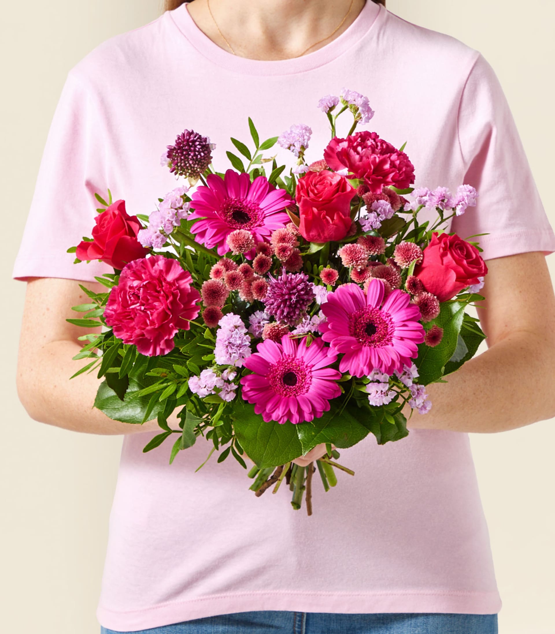 Eine Person in einem rosa T-Shirt hält einen farbenfrohen Strauß rosa und roter Blumen vor einem schlichten Hintergrund.