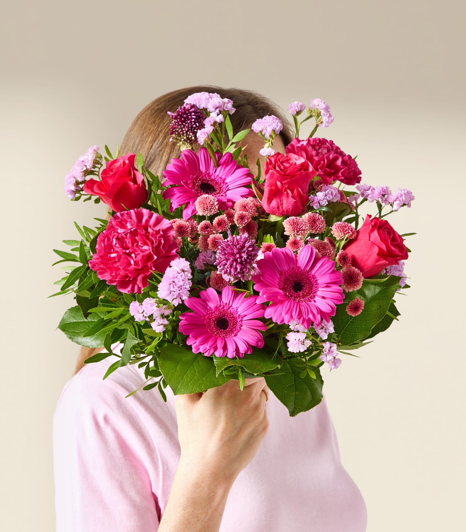 Eine Person in einem rosa Hemd hält einen leuchtenden Strauß rosa und roter Blumen, der ihr Gesicht vor einem schlichten, hellen Hintergrund verdeckt.