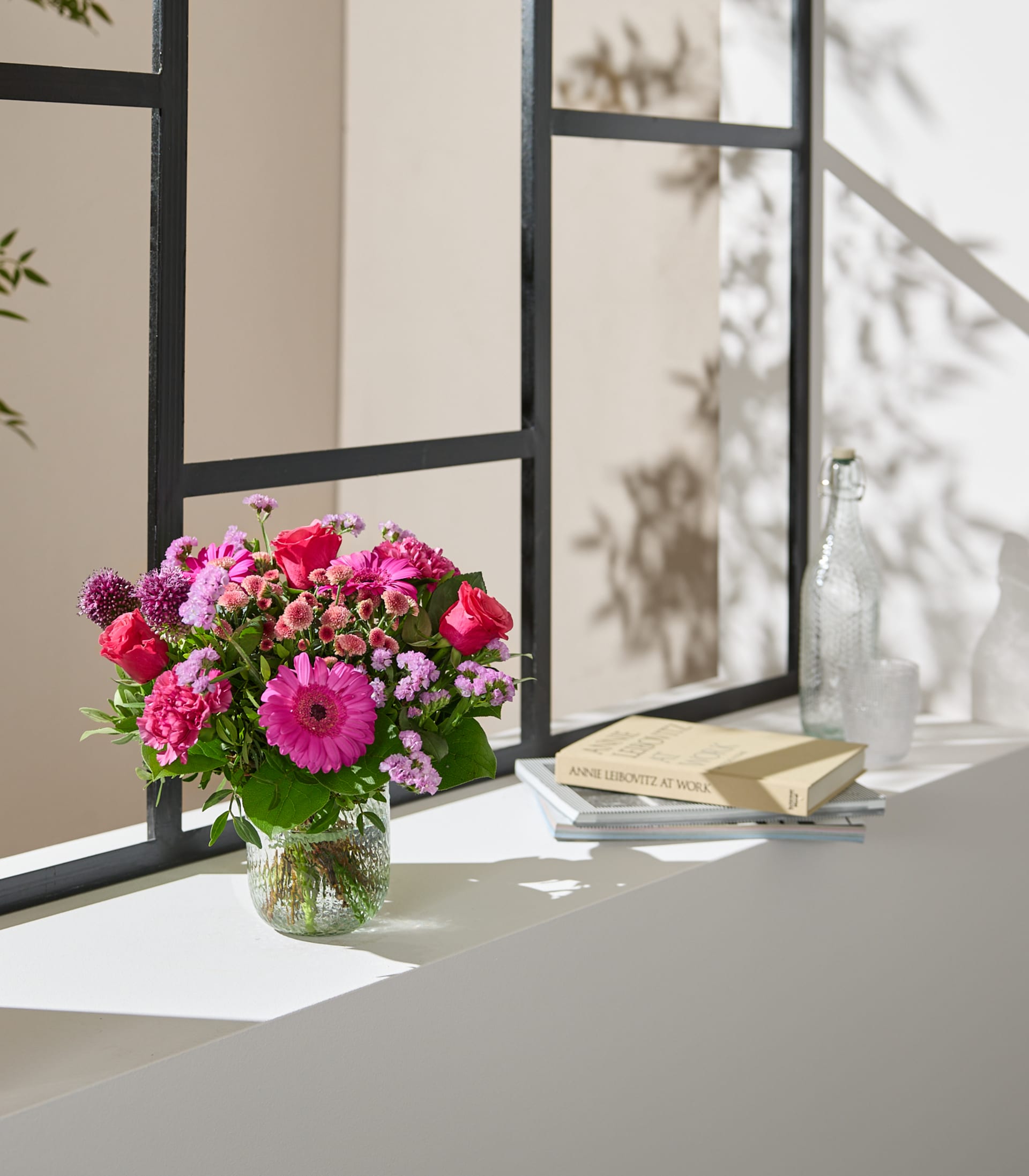 Auf einem Fensterbrett steht eine Vase mit leuchtend pinken und roten Blumen; im Hintergrund sind vor einer neutral getönten Wand und gerahmten Fenstern ein Buch und eine Pflanze zu sehen.