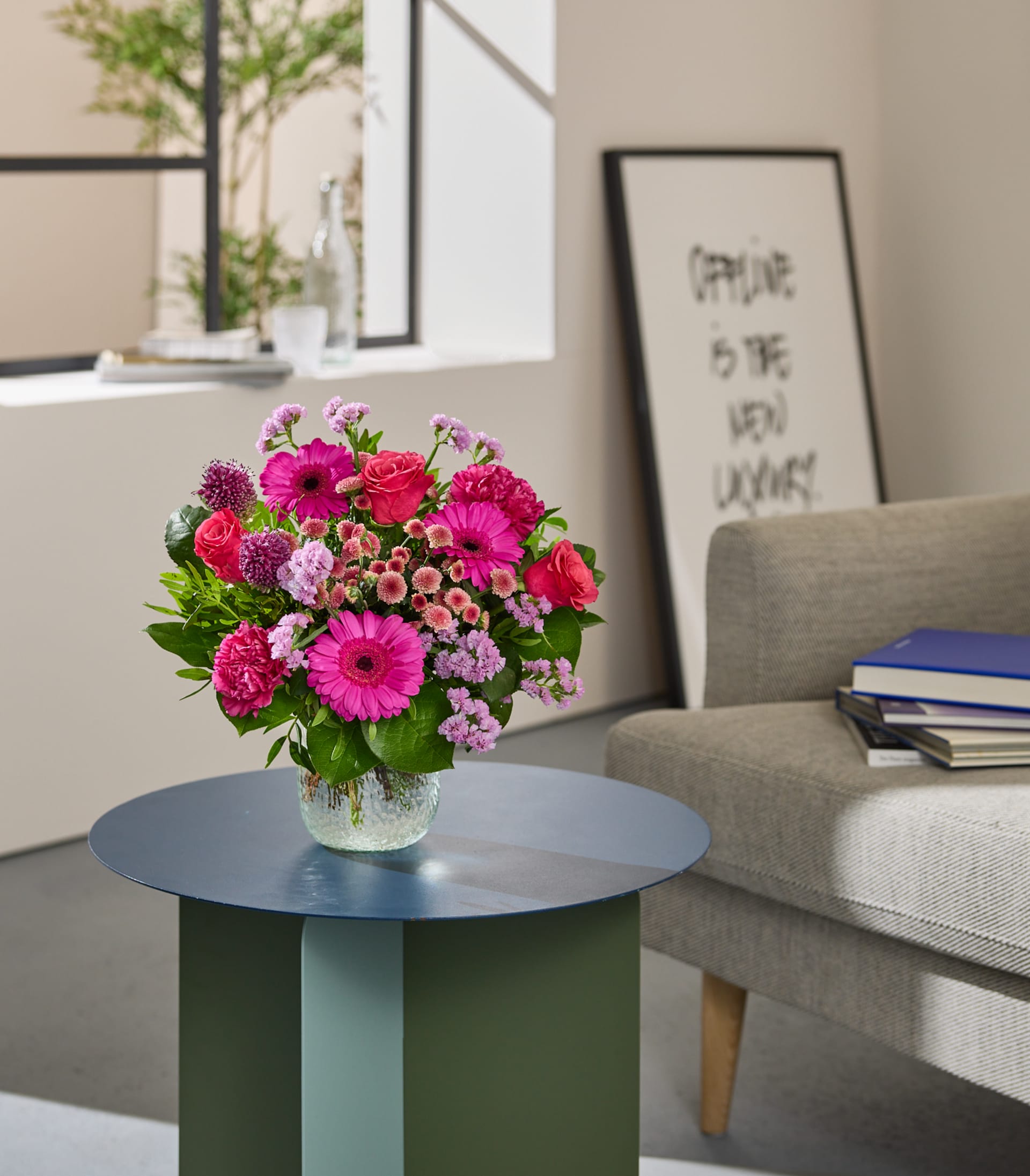 Auf einem kleinen runden Tisch in einem gemütlichen Wohnzimmer steht ein farbenfroher Strauß rosa und lila Blumen; im Hintergrund sind ein gerahmtes Kunstwerk und ein Bücherstapel zu sehen.