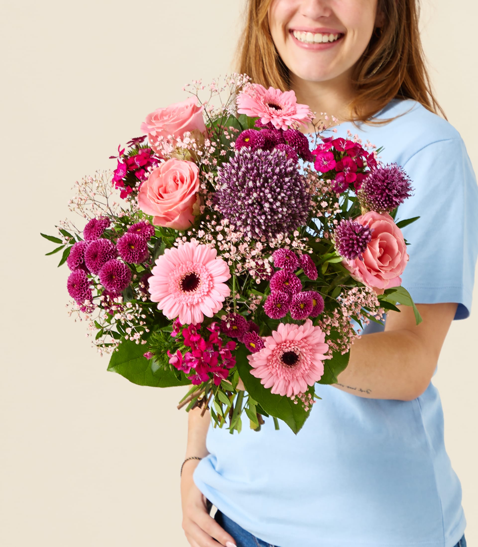 Eine lächelnde Person in einem blauen Hemd hält einen farbenfrohen Strauß rosa und lila Blumen vor einem schlichten, hellen Hintergrund.