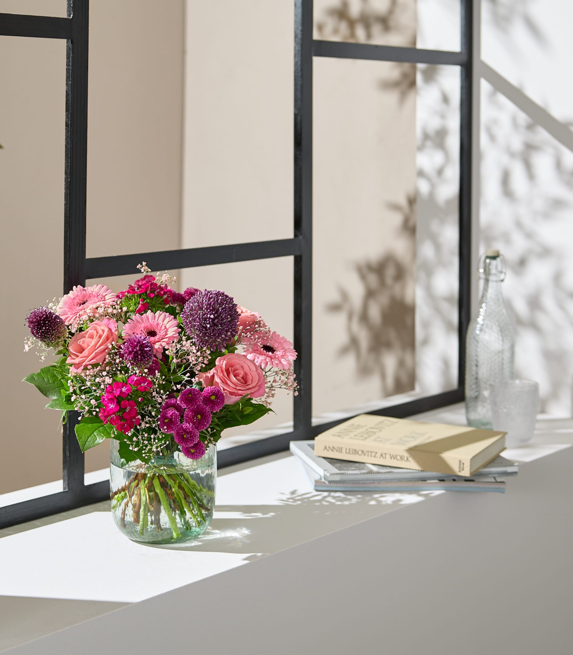 Auf einem Fensterbrett steht ein farbenfroher Strauß rosa und violetter Blumen, im Hintergrund ein Buch und andere Gegenstände, die eine gemütliche und einladende Szene schaffen.