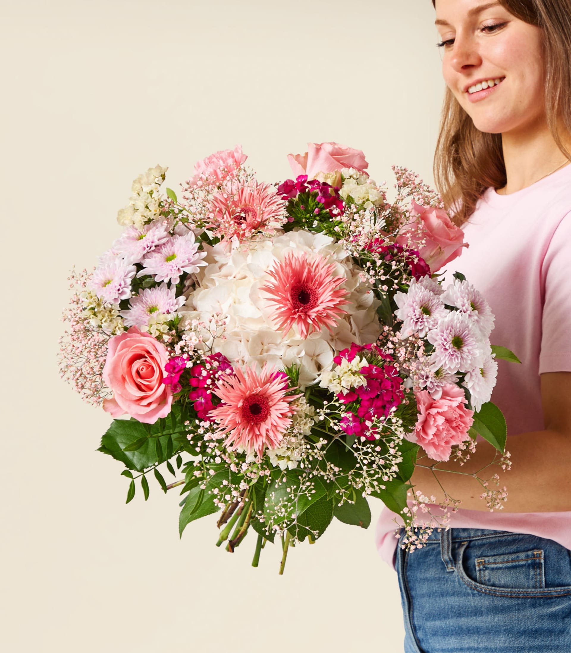 Eine Frau in einem rosa Hemd und Jeans hält einen großen Strauß rosa und weißer Blumen vor einem schlichten, hellen Hintergrund.