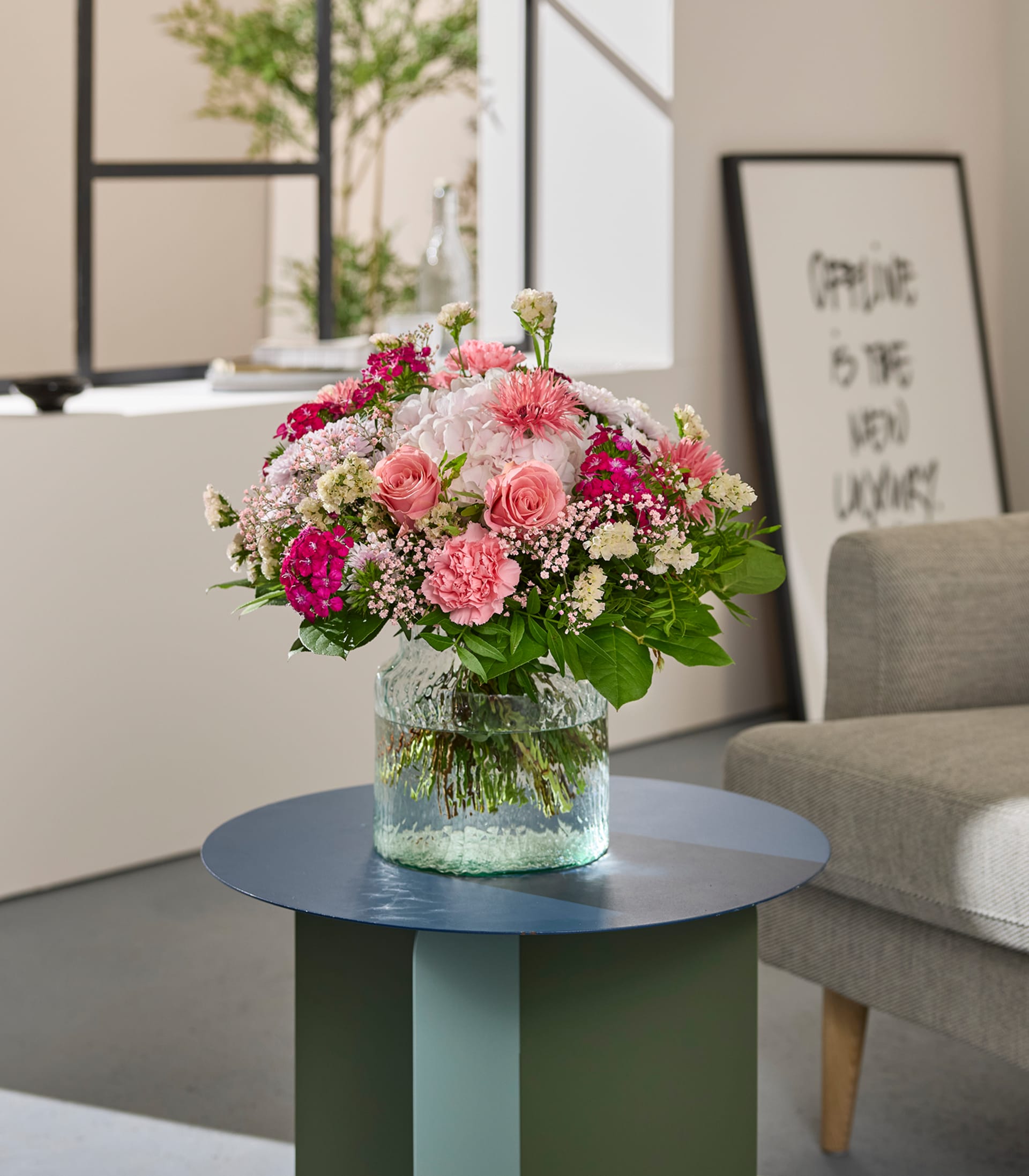 Auf einem modernen Beistelltisch steht ein farbenfroher Strauß rosa und weißer Blumen in einer Glasvase; im sanft beleuchteten Hintergrund sind ein gerahmtes abstraktes Kunstwerk und ein bequemer Sessel zu sehen.