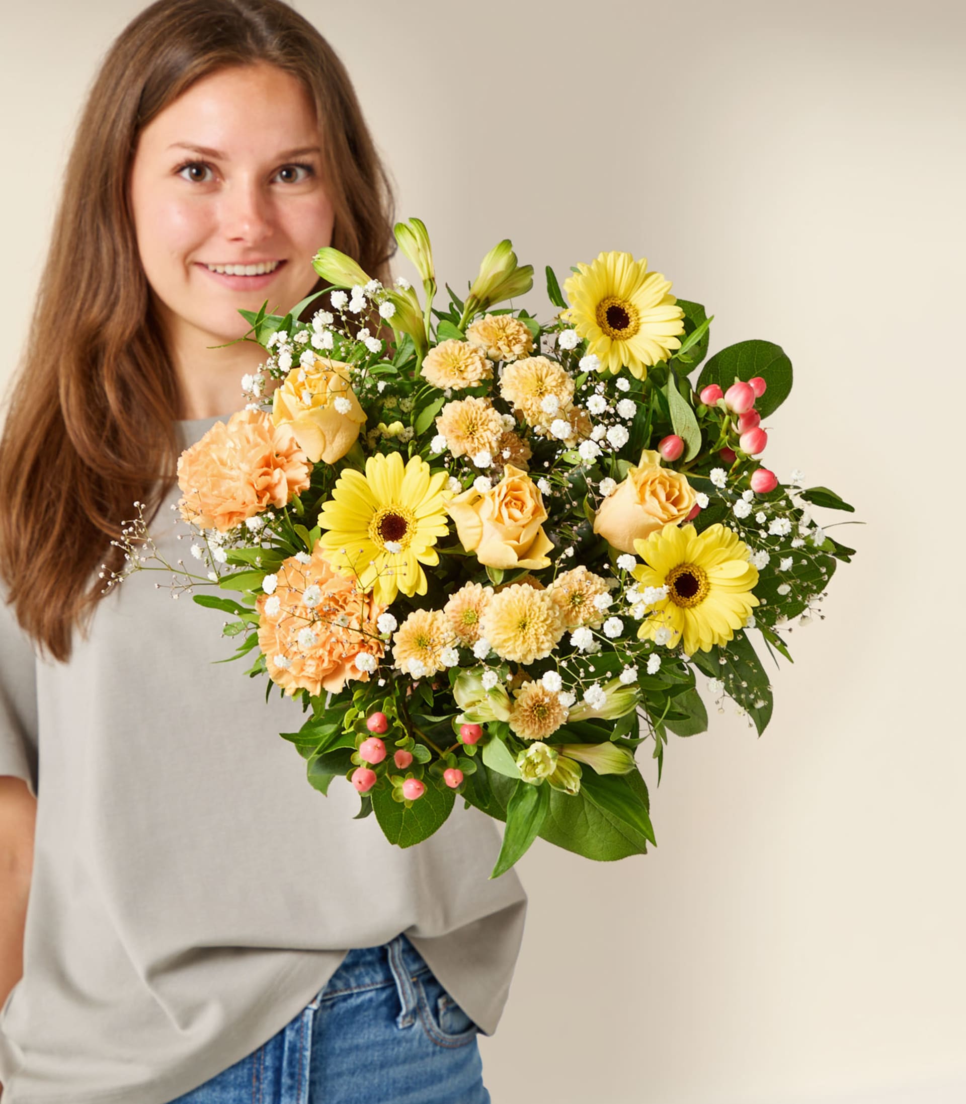 Eine lächelnde Frau in grauem Hemd und blauen Jeans hält einen leuchtenden Strauß gelber und orangefarbener Blumen vor einem schlichten, hellen Hintergrund.