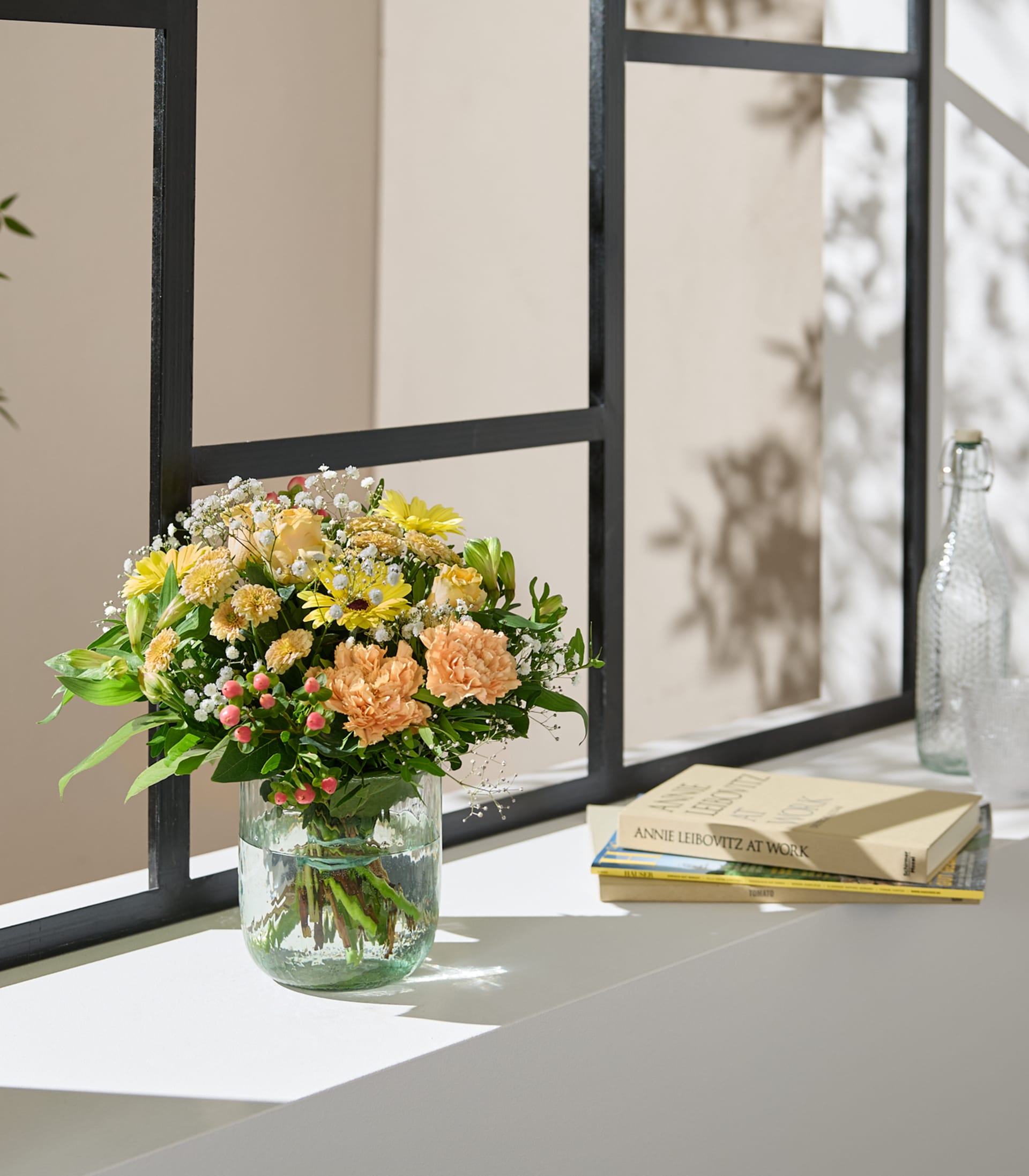 Eine Hand hält einen leuchtenden Strauß gelber und orangefarbener Blumen mit zarten weißen Füllblumen vor einem sanften, neutralen Hintergrund.