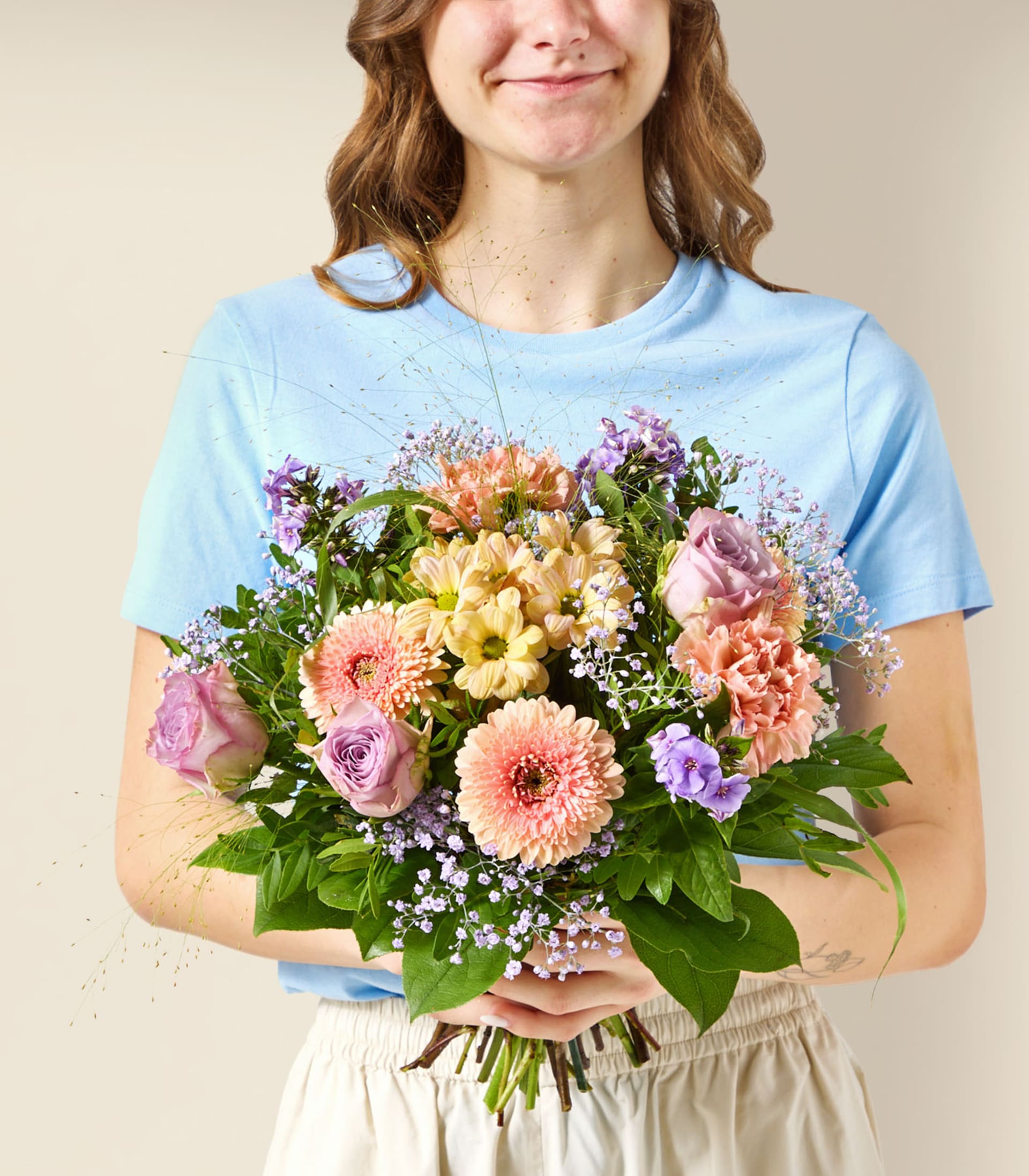 Eine junge Frau in einem blauen T-Shirt und weißen Shorts hält einen farbenfrohen Strauß rosa, lila und gelber Blumen vor einem schlichten, hellen Hintergrund.