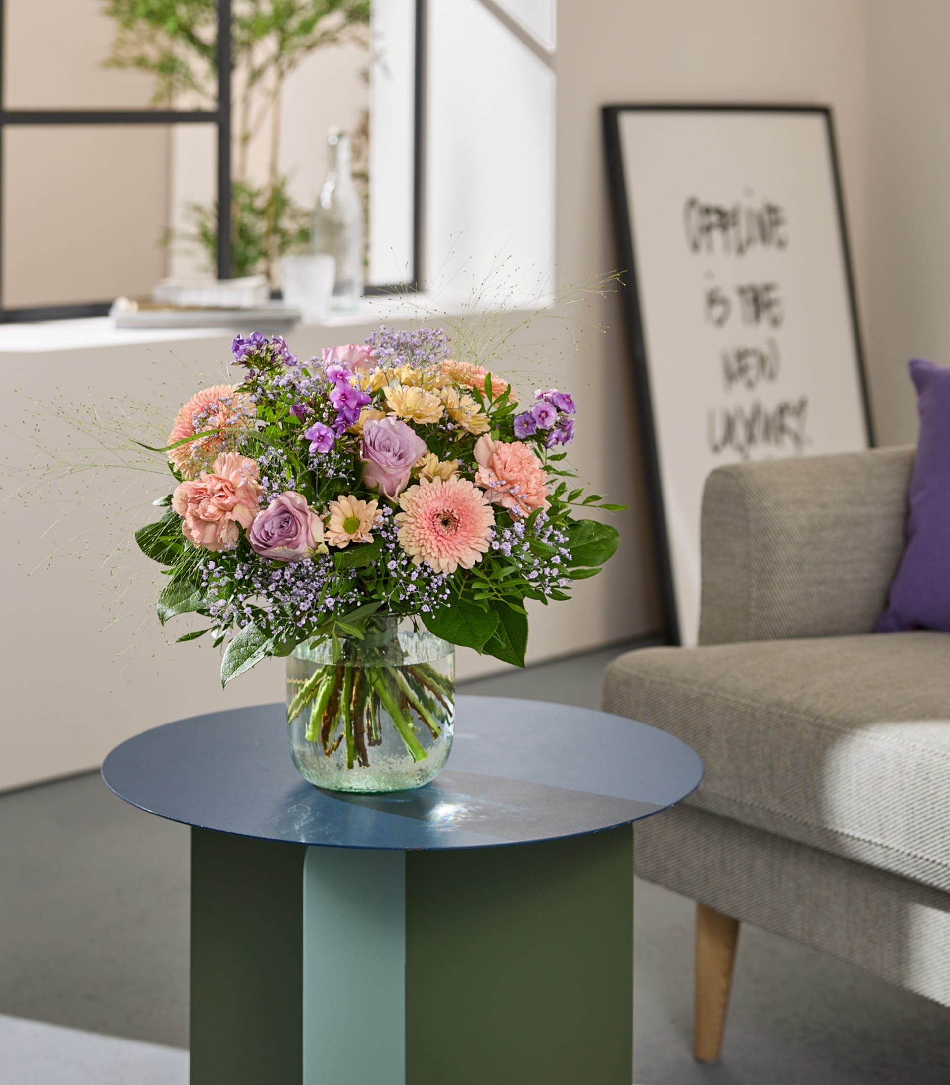 Ein leuchtender Strauß rosa und violetter Blumen steht in einer Glasvase auf einem Fensterbrett, im Hintergrund sind Bücher und eine Flasche zu sehen.