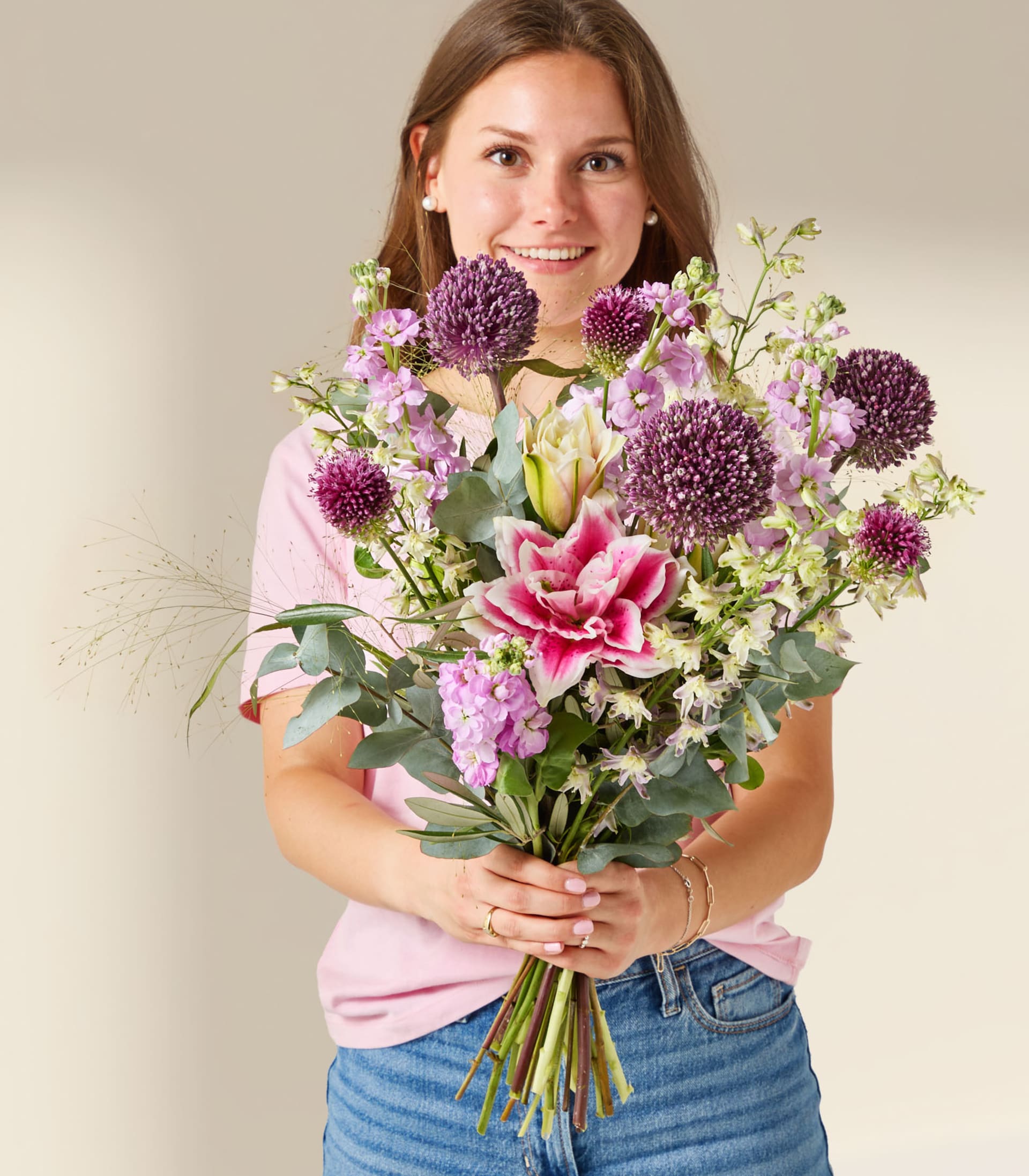 Eine lächelnde junge Frau in einem rosa T-Shirt und Jeansshorts hält vor einem schlichten, hellen Hintergrund einen großen Strauß lila und rosa Blumen.