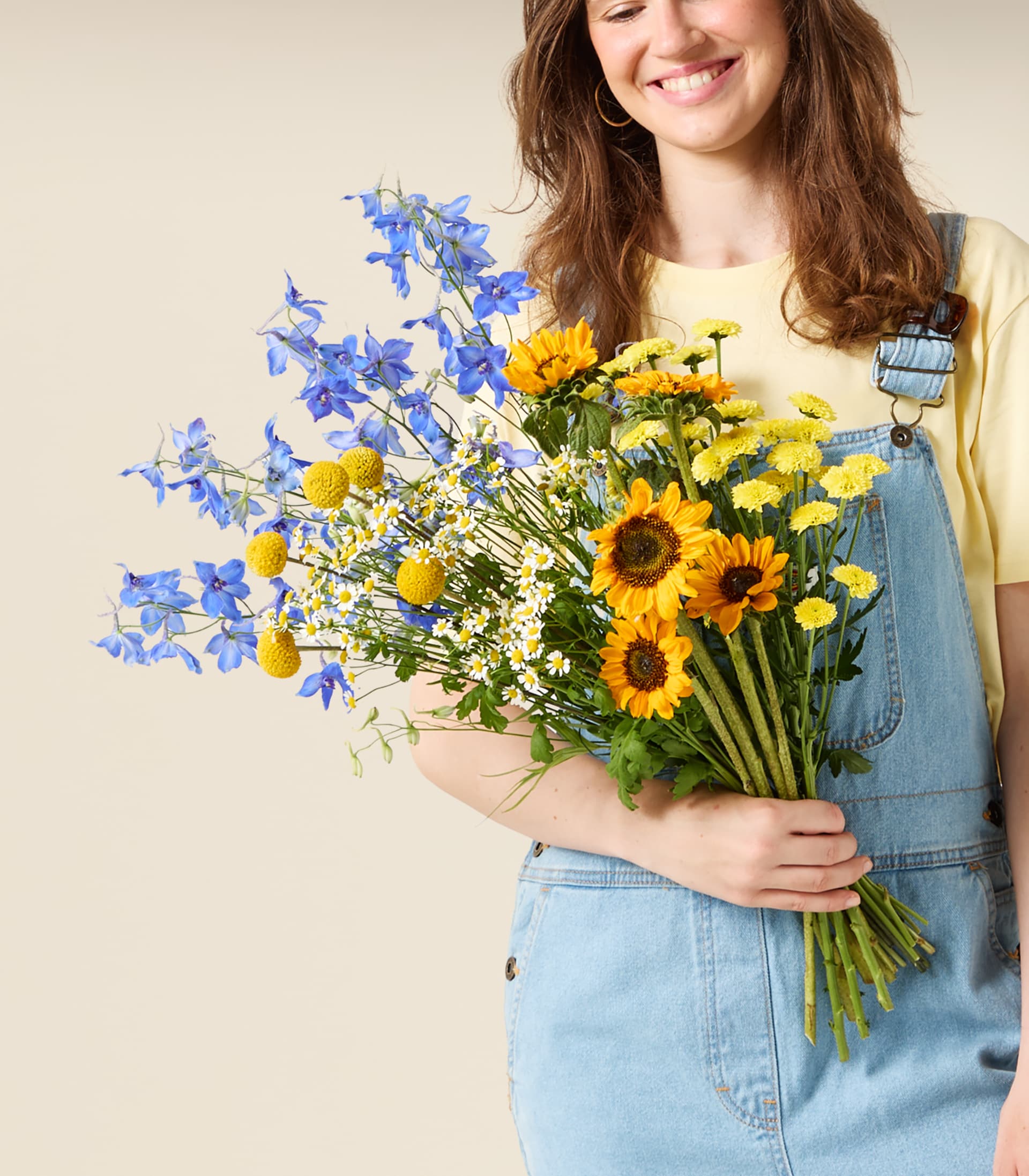 Eine lächelnde Frau in Jeanslatzhose hält einen farbenfrohen Strauß aus blauen Ritterspornen, gelben Sonnenblumen und kleinen weißen und gelben Blüten vor einem schlichten, hellen Hintergrund.
