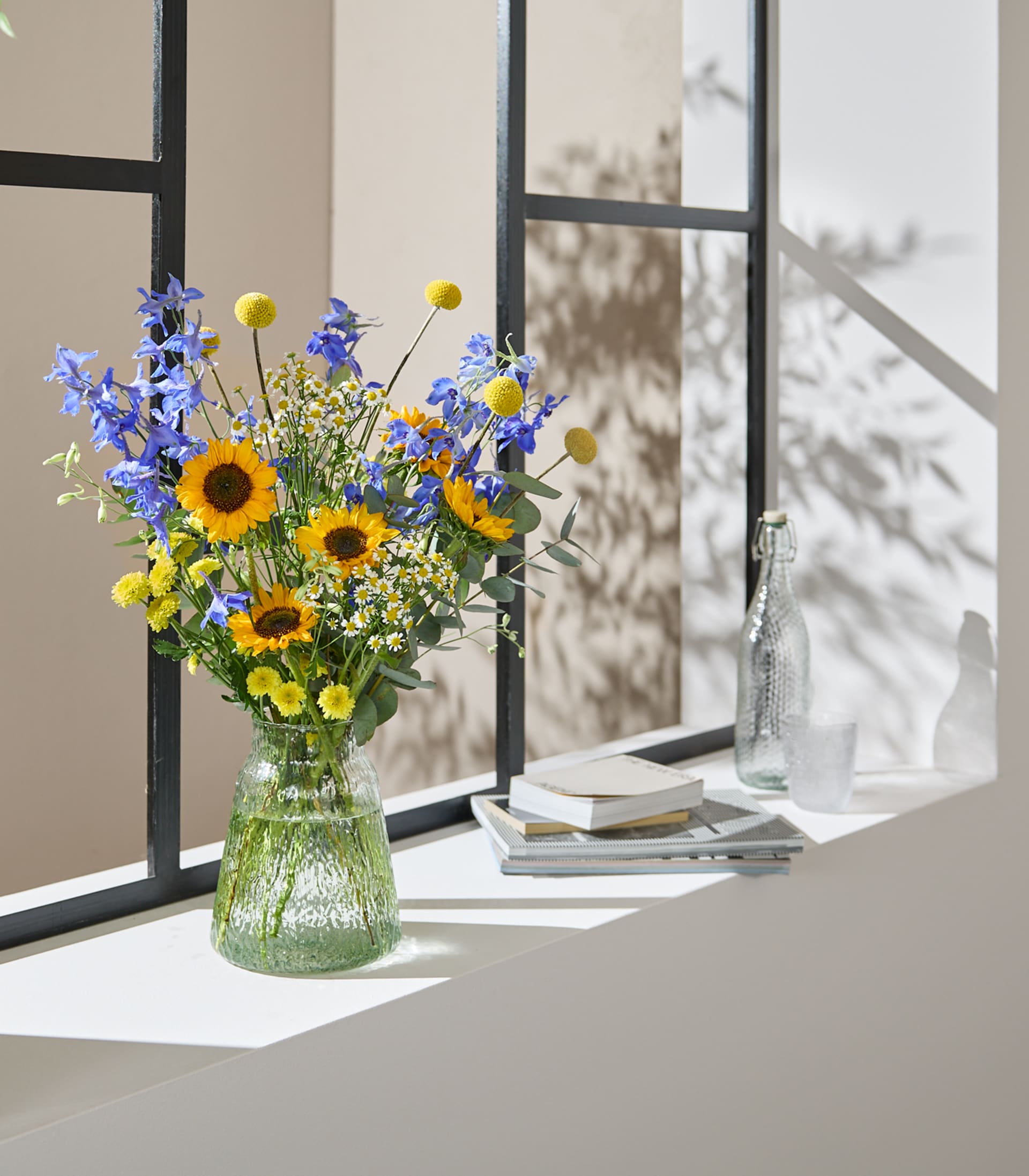 Ein leuchtender Strauß Sonnenblumen und blauer Rittersporne steht in einer strukturierten grünen Vase auf einem Fensterbrett, im Hintergrund werfen Bücher und eine Glasflasche gefleckte Schatten.