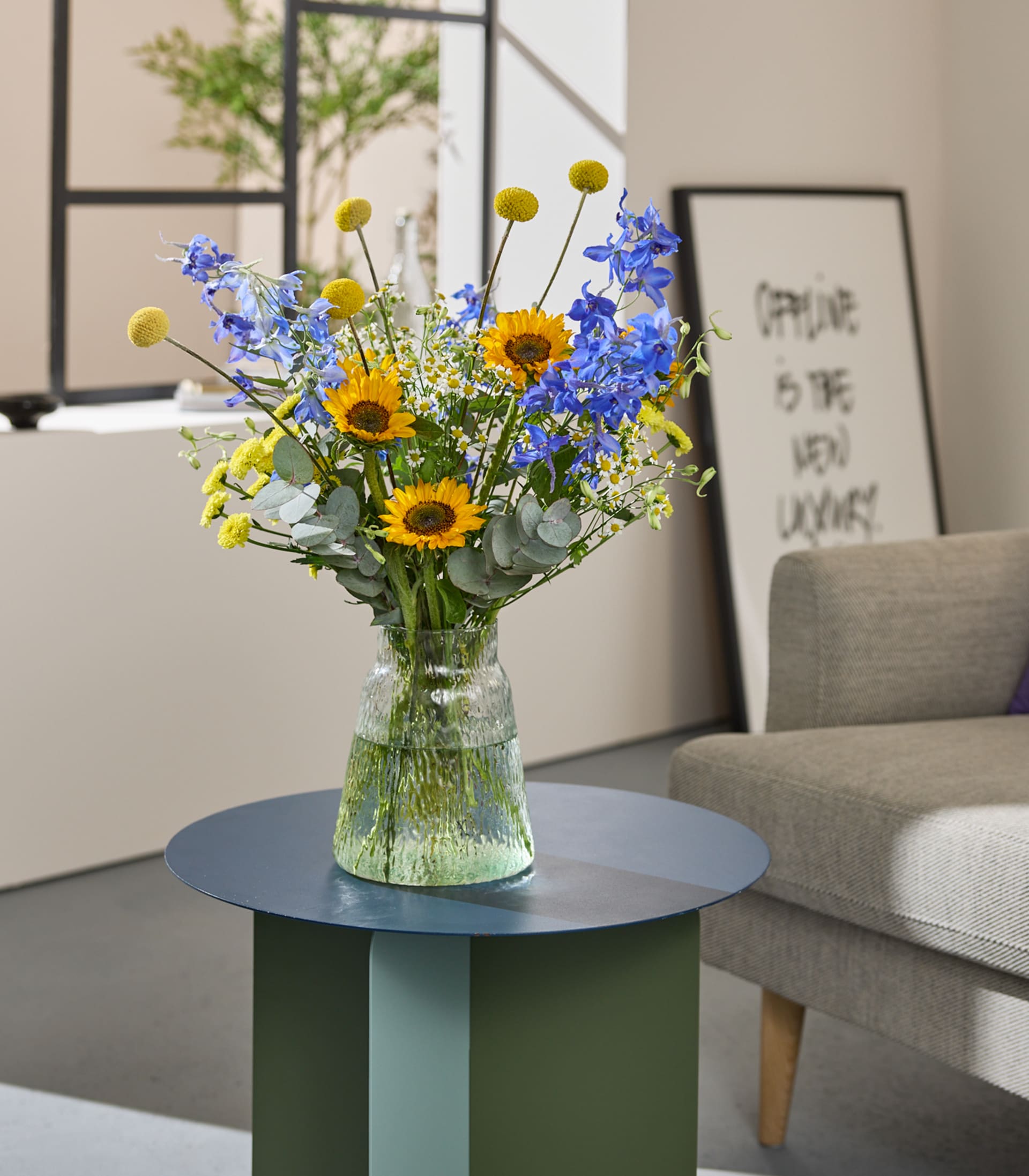 Ein leuchtender Strauß Sonnenblumen und blauer Rittersporne steht in einer Glasvase auf einem grünen Tisch, im Hintergrund ein gerahmter abstrakter Druck und ein graues Sofa.