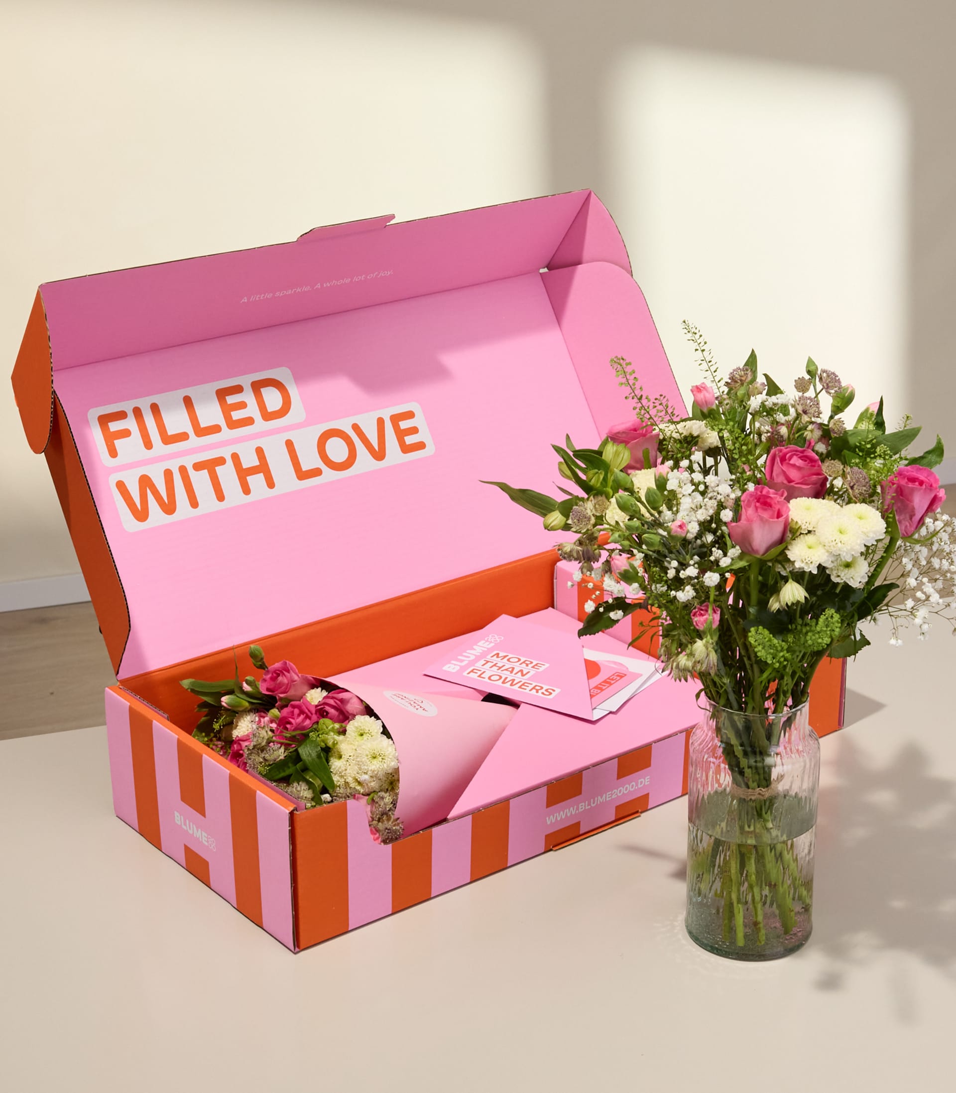Auf einem Tisch steht eine leuchtend pink-orangefarbene Geschenkbox mit der Aufschrift „Mit Liebe gefüllt“, daneben eine Vase mit bunten Blumen.