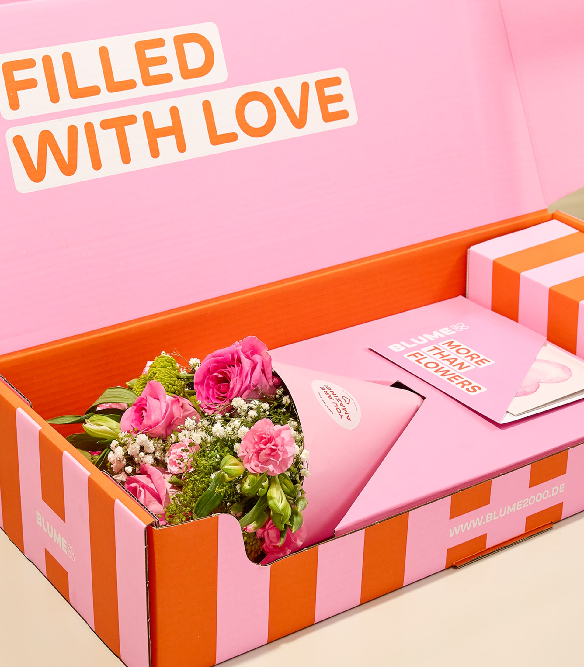 Eine leuchtend pink-orange gestreifte Schachtel enthält einen Strauß rosa Rosen und Grünzeug, begleitet von einer Karte mit dem Text „Mit Liebe gefüllt“ auf zartrosa Hintergrund.