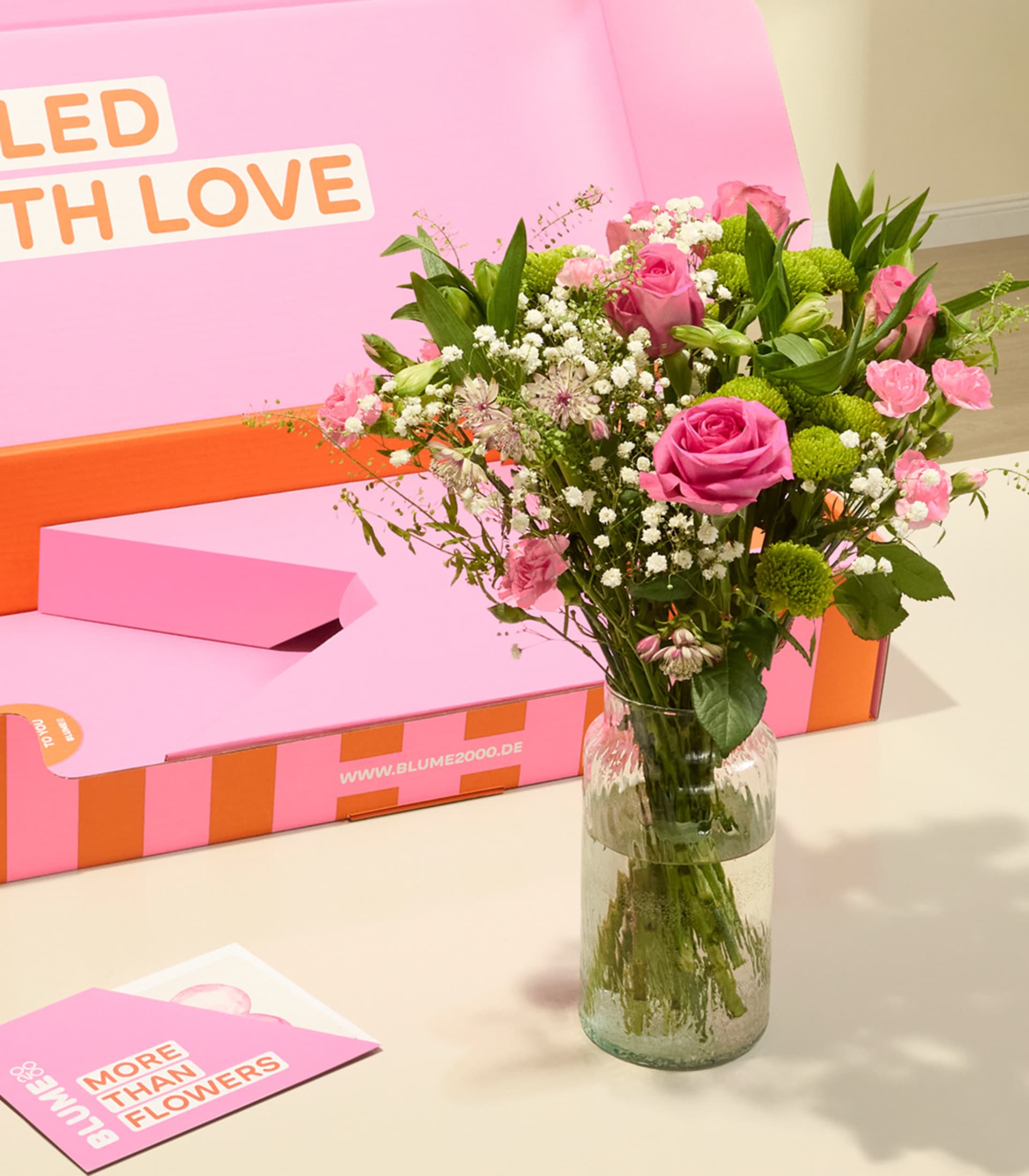 Auf einem Tisch steht ein Strauß rosa Rosen und anderer Blumen in einer Glasvase; im Hintergrund ist eine rosa-orangefarbene Geschenkbox mit der Aufschrift „Mit Liebe gefüllt“ zu sehen.