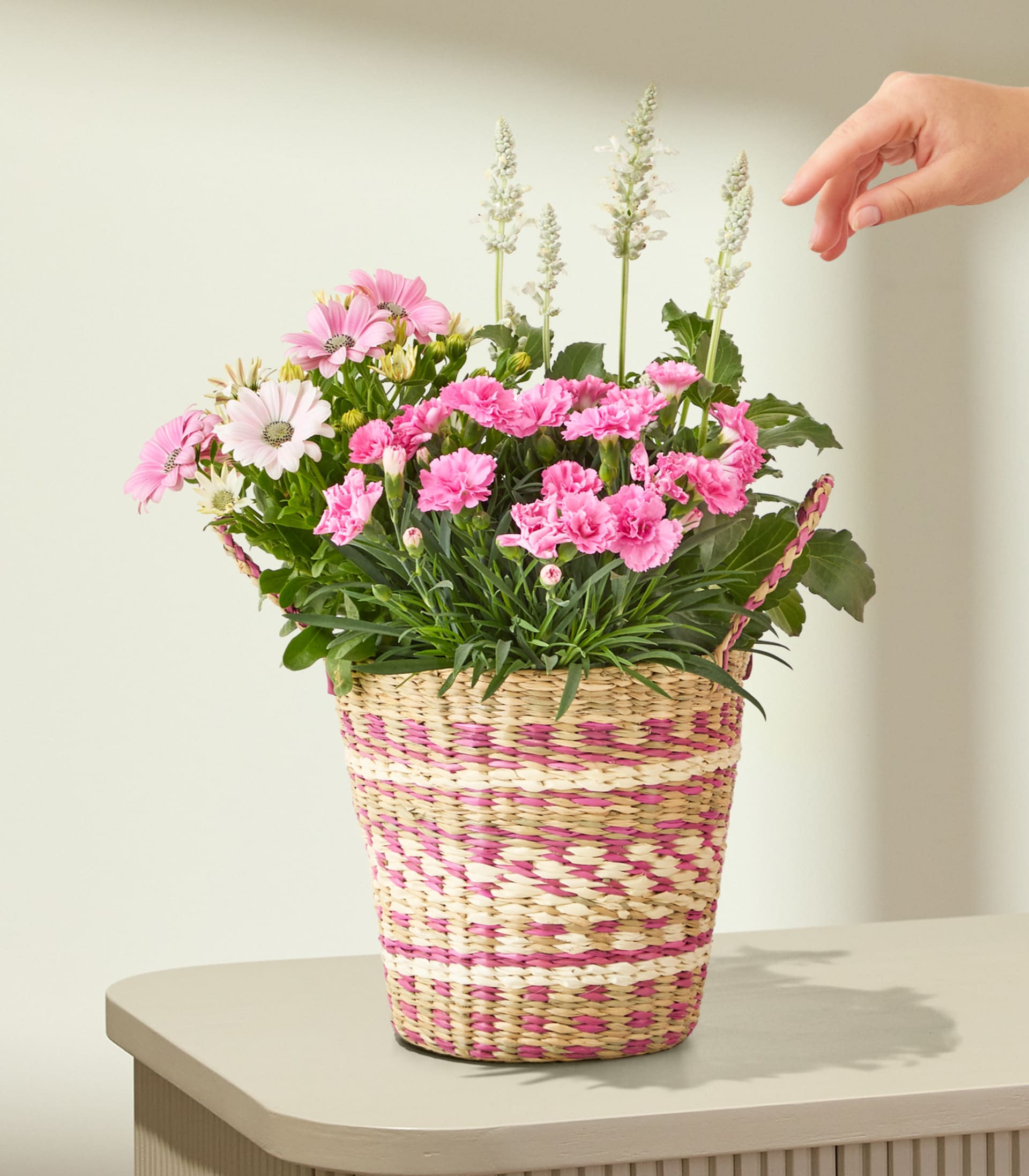 Auf einem Tisch steht ein geflochtener Korb, gefüllt mit rosa und weißen Blumen; eine Hand streckt sich vor einer hellen Wand danach aus.