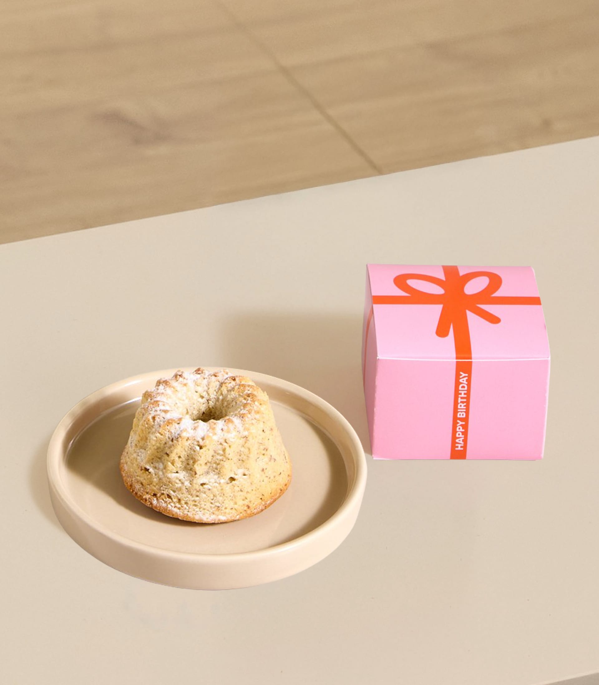 Auf einem Teller neben einer rosa Geschenkbox auf hellem Untergrund steht ein Gugelhupf; im Hintergrund ist ein verschwommener Holzboden zu sehen.