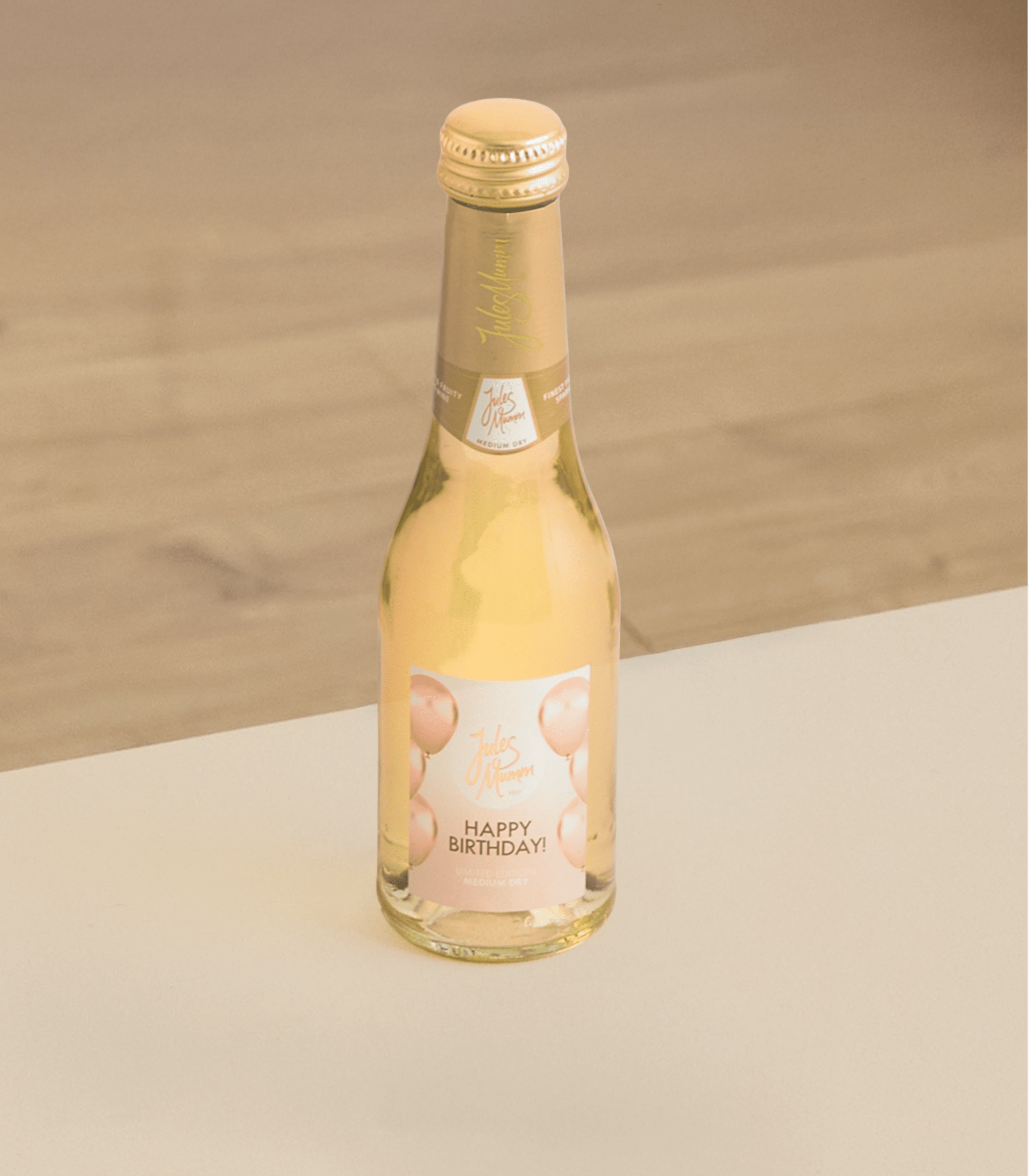 Auf einer weißen Fläche steht eine kleine Flasche Apfelschaumwein mit der Aufschrift „Alles Gute zum Geburtstag!“, der Hintergrund ist verschwommen.
