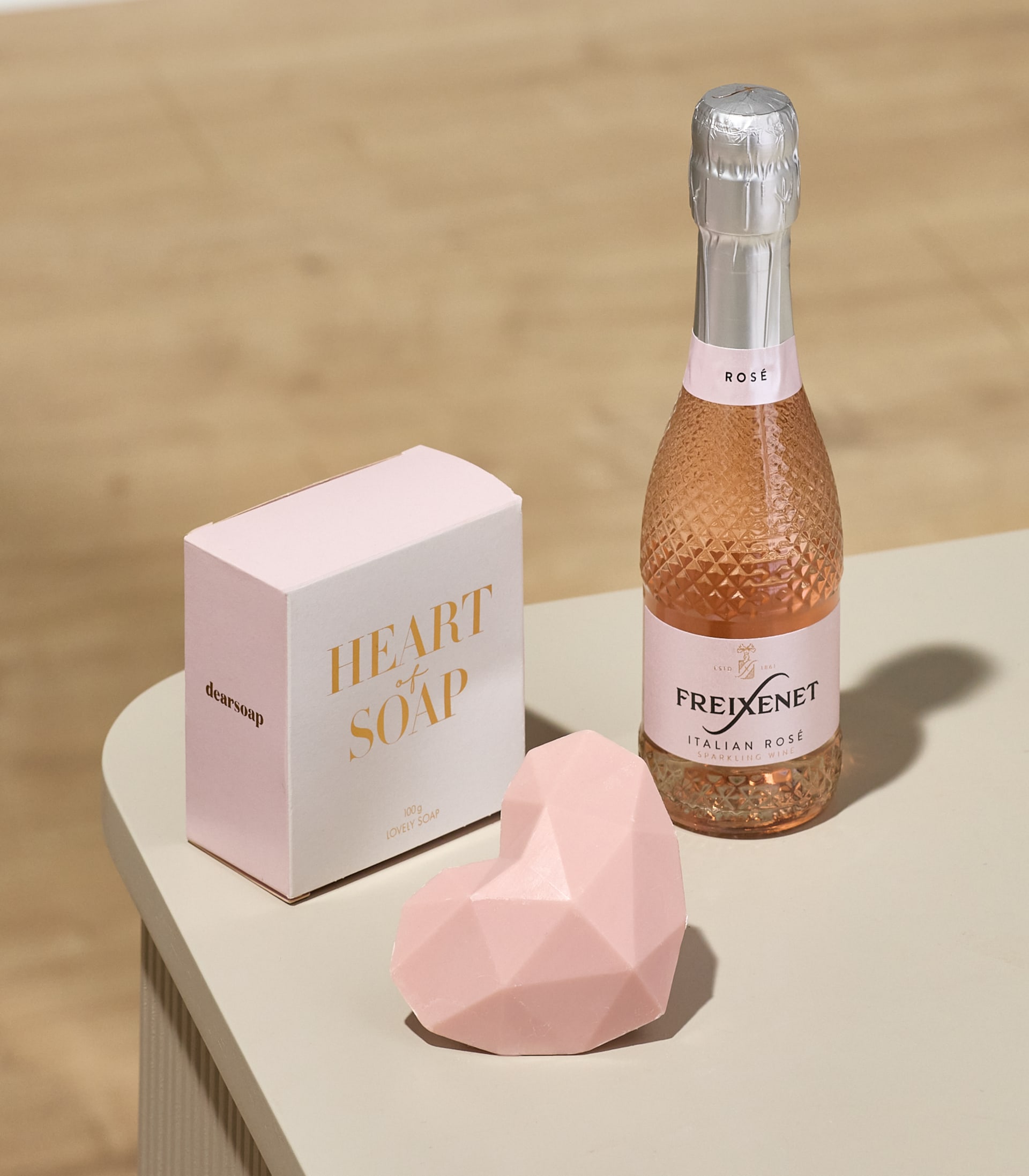 Im Vordergrund stehen eine rosafarbene, geometrische, herzförmige Seife und eine Schachtel mit der Aufschrift „HERZ DER SEIFE“ auf einer hellrosa Fläche, im Hintergrund eine kleine Flasche Roséwein vor einer hellen Holzmaserung.