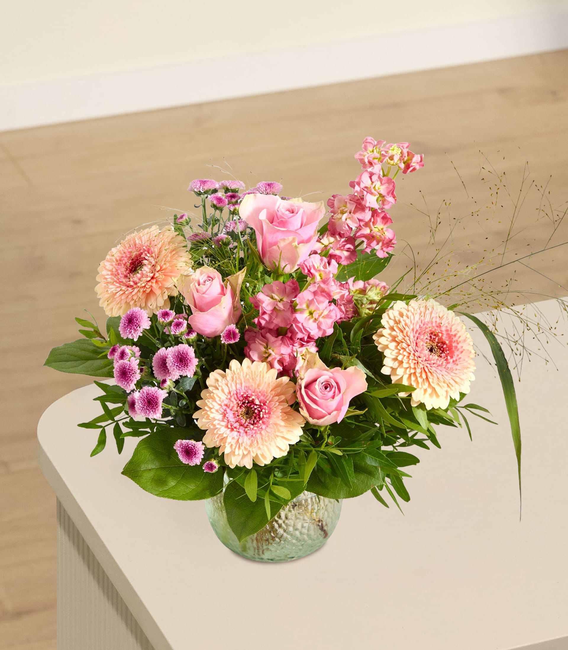 Ein zarter Strauß rosa und pfirsichfarbener Blüten steht in einer strukturierten Glasvase auf einer hellen Oberfläche, im Hintergrund sind Wand und Boden sanft verschwommen zu sehen.