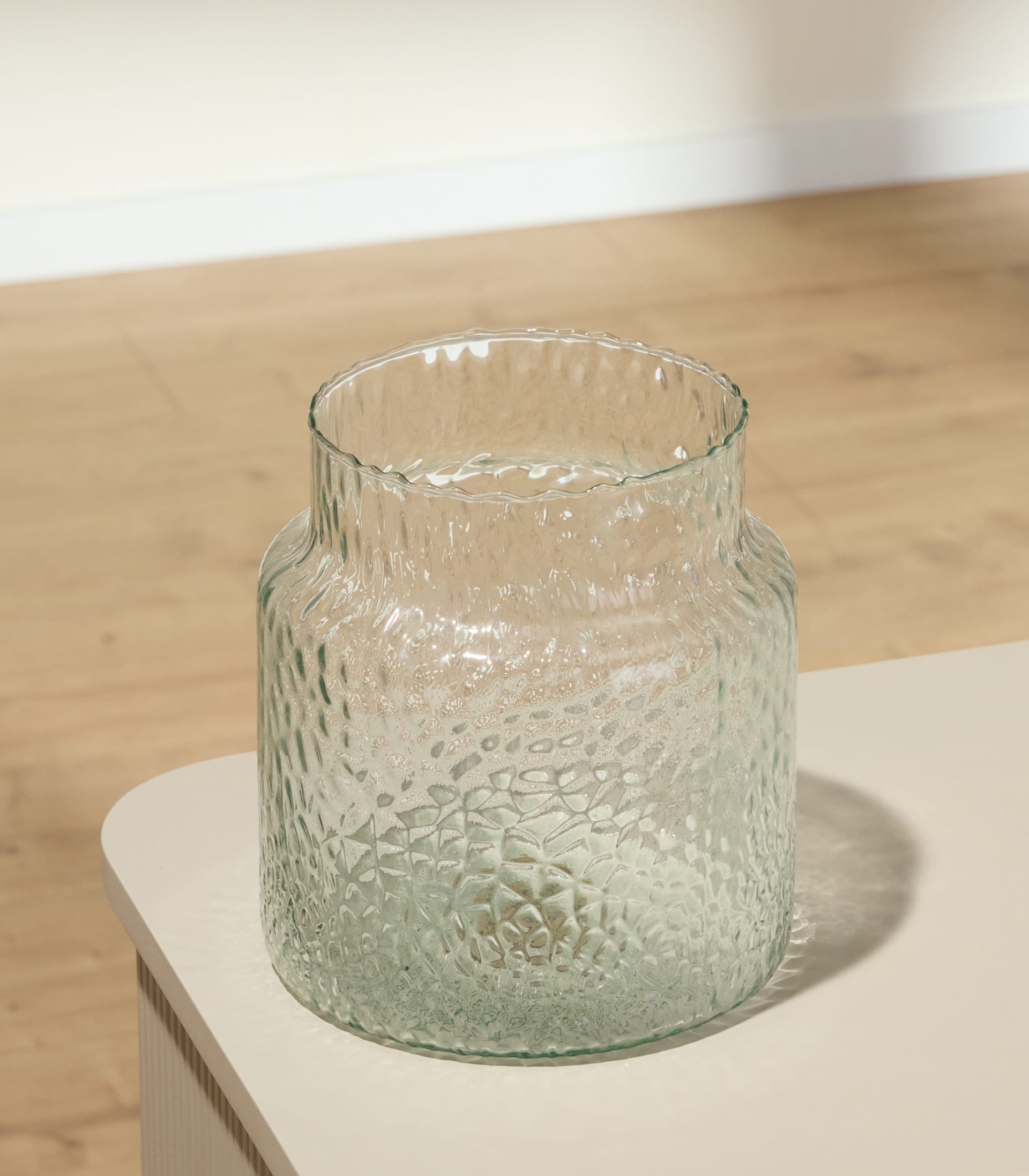 Im Vordergrund steht eine strukturierte Glasvase auf einer hellen Fläche; im Hintergrund sind ein Holzboden und eine weiße Wand zu sehen.