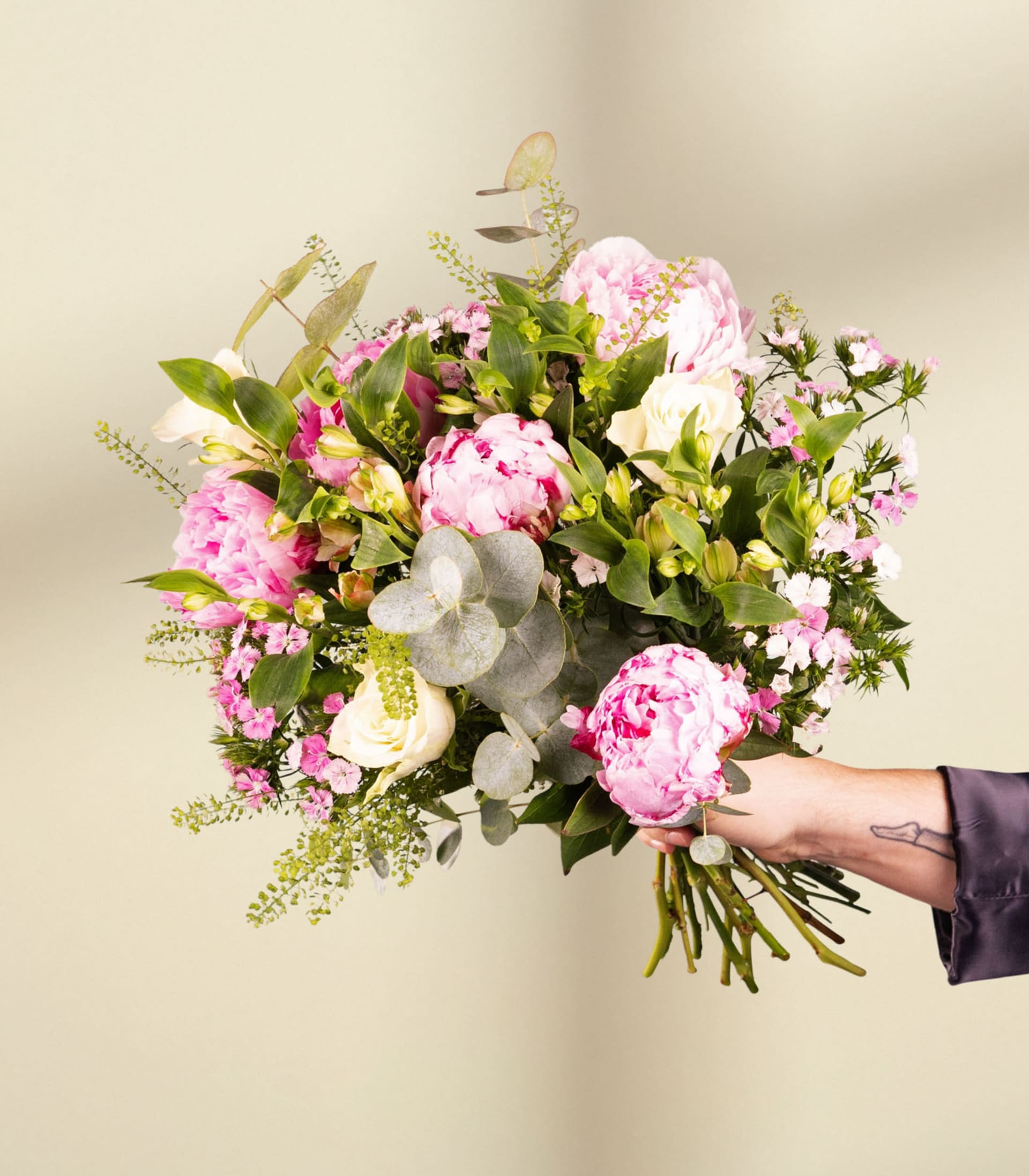 Ein lebendiger Strauß rosa und weißer Blumen, darunter üppige Pfingstrosen, eingebettet zwischen grünem Laub vor einem neutralen Hintergrund.