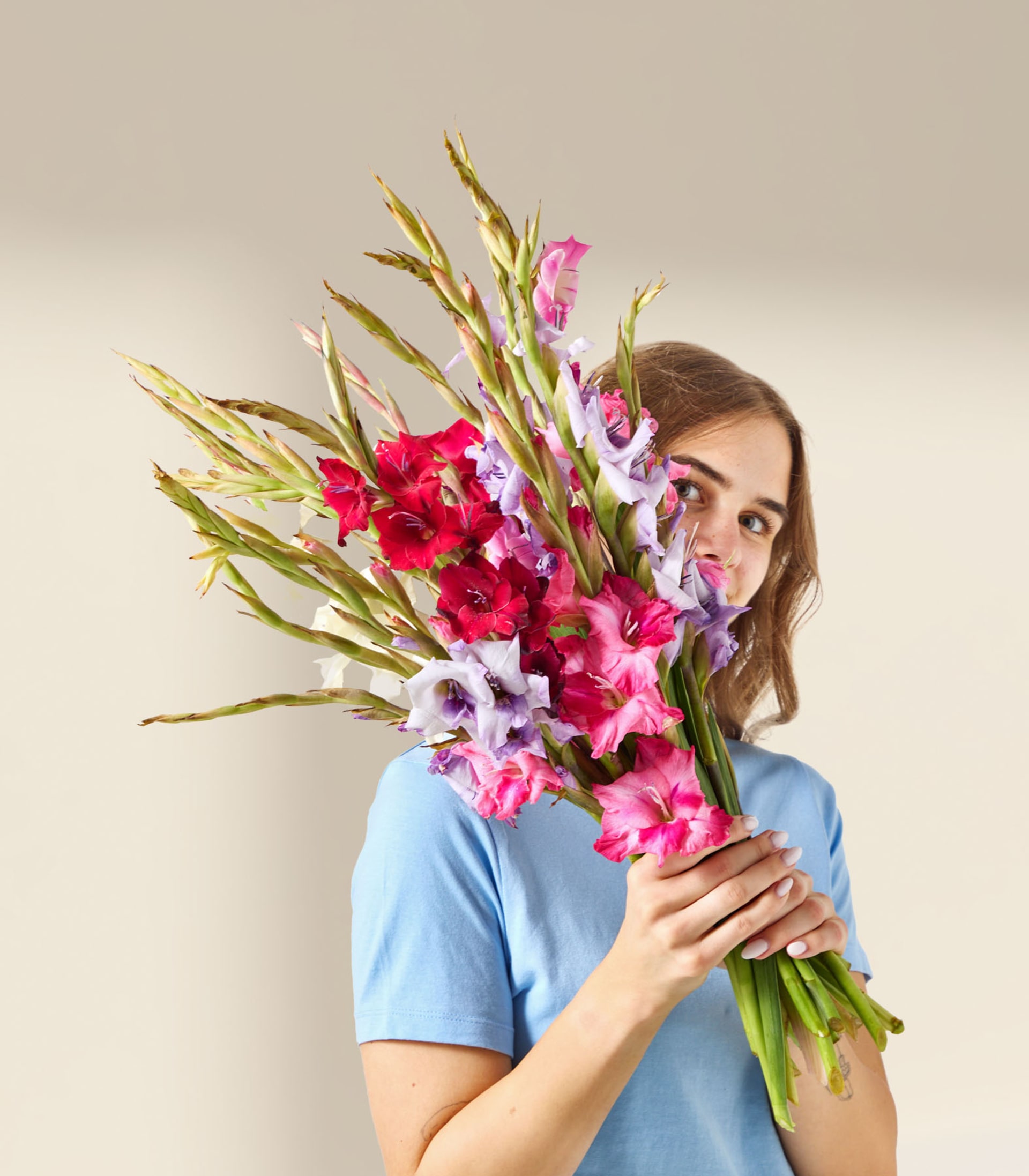 Ein leuchtender Strauß bunter Blumen, darunter rote und rosa Gladiolen, wird vor den Körper einer Person gehalten und verdeckt so ihr Gesicht.