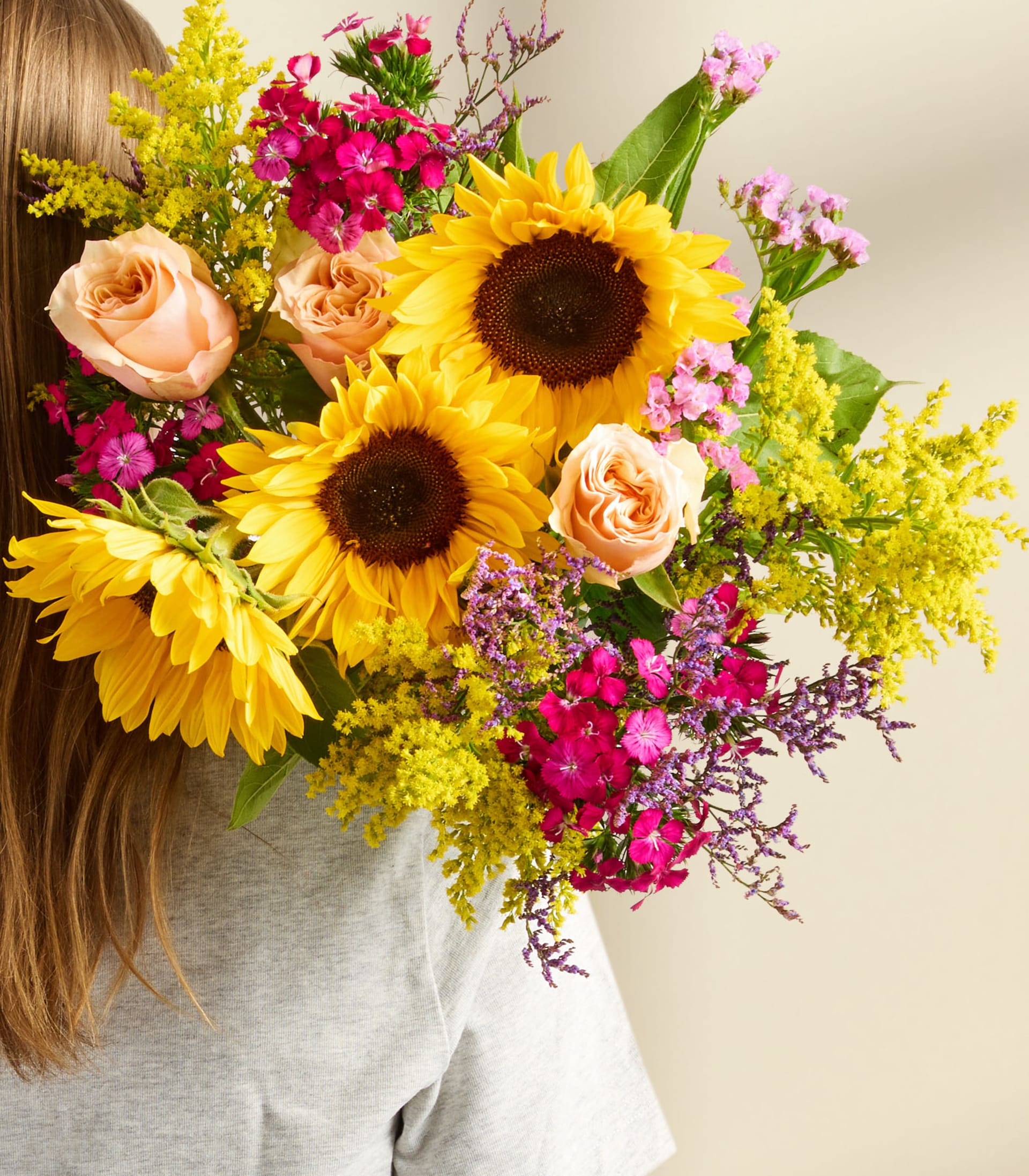 Ein leuchtender Strauß aus Sonnenblumen, Rosen und anderen bunten Blumen wird vor das Gesicht einer Person gehalten und verdeckt so ihre Gesichtszüge.