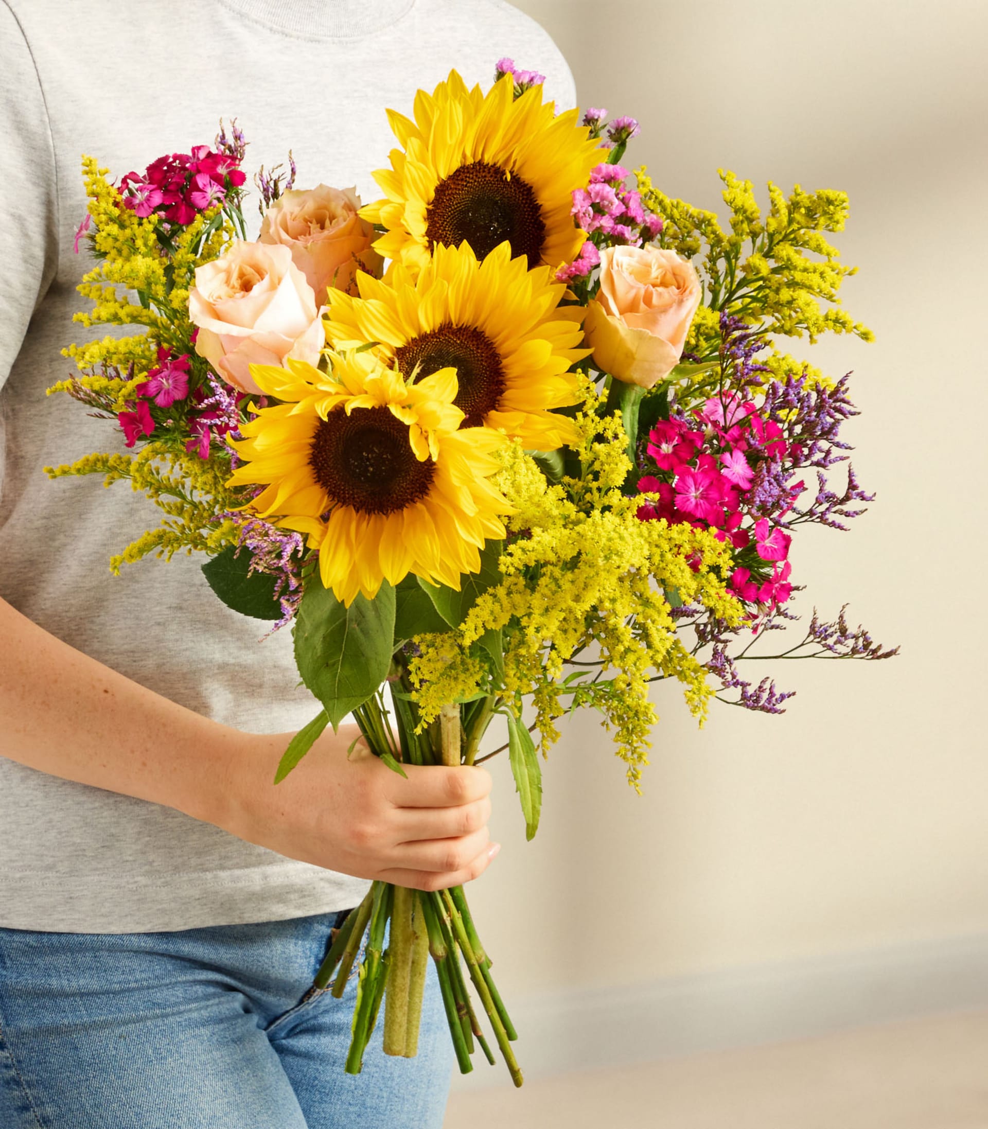 Ein lebendiger Strauß aus Sonnenblumen, Rosen und anderen bunten Blumen, arrangiert in einer Glasvase vor einem schlichten weißen Hintergrund.