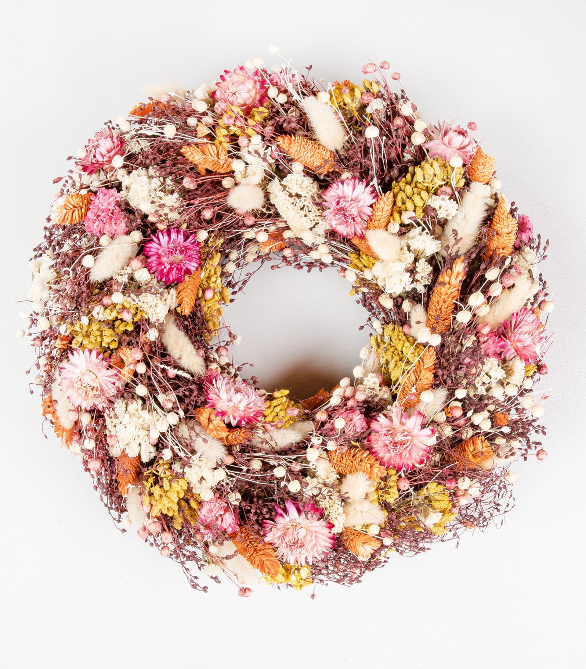 Ein lebendiger, runder Kranz aus verschiedenen getrockneten Blumen und Pflanzen, darunter rosa, gelbe und weiße Blüten sowie Zweige und andere natürliche Elemente, vor einem schlichten weißen Hintergrund.