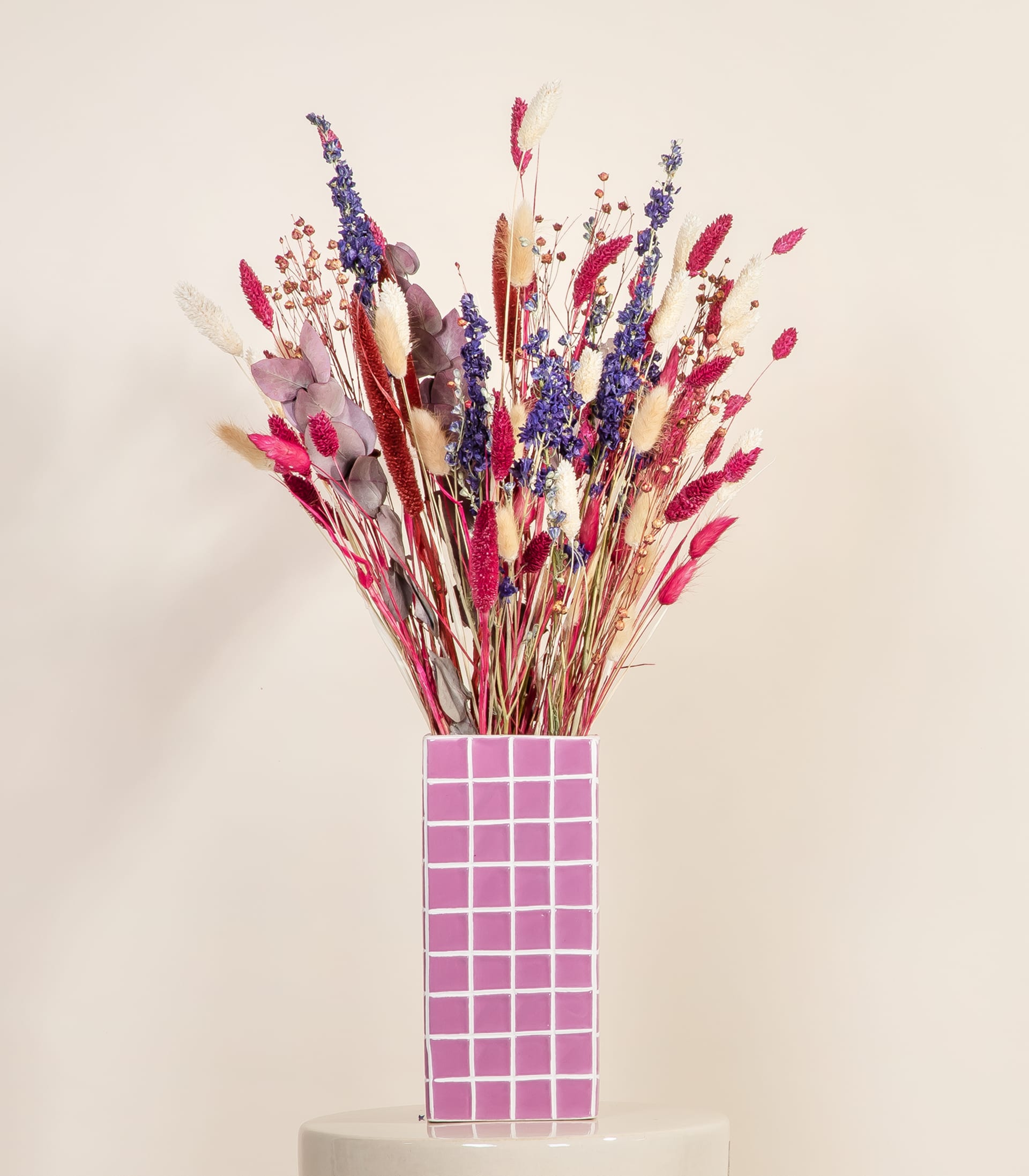 Ein farbenfroher Strauß getrockneter Blumen und Federn entspringt einer rosa Vase mit Gittermuster vor einem schlichten, hellen Hintergrund.