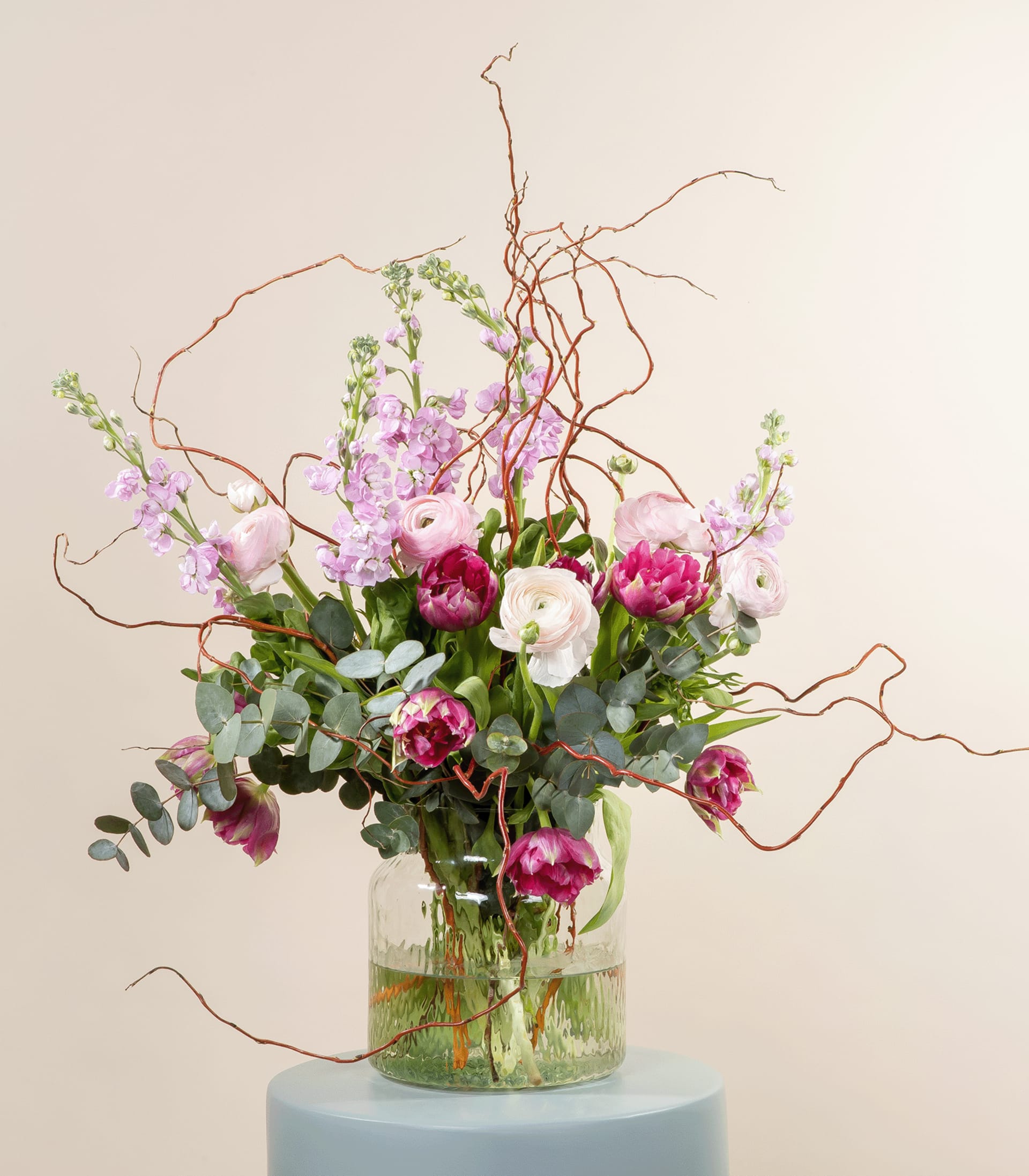 Ein farbenfroher Strauß rosa und weißer Blumen, darunter Pfingstrosen und andere Blüten, arrangiert in einer Glasvase mit Zweigen und Grünpflanzen vor einem schlichten, hellen Hintergrund.