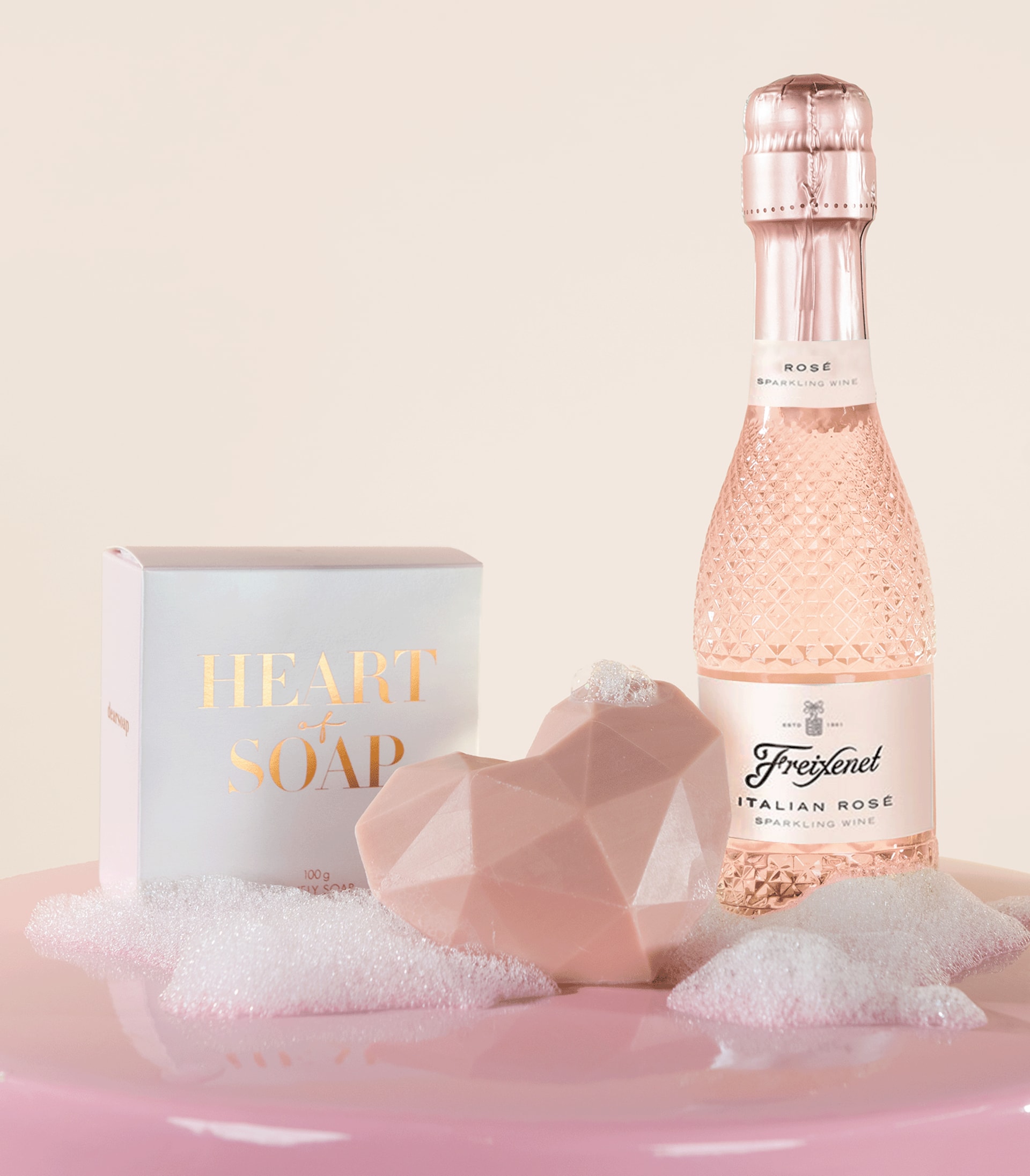 Eine rosa getönte Glasflasche Sekt, eine weiße Schachtel mit der Aufschrift „Luxusseife“ und ein rosafarbener, geometrisch geformter Gegenstand stehen auf einer weichen, flauschigen rosafarbenen Oberfläche.