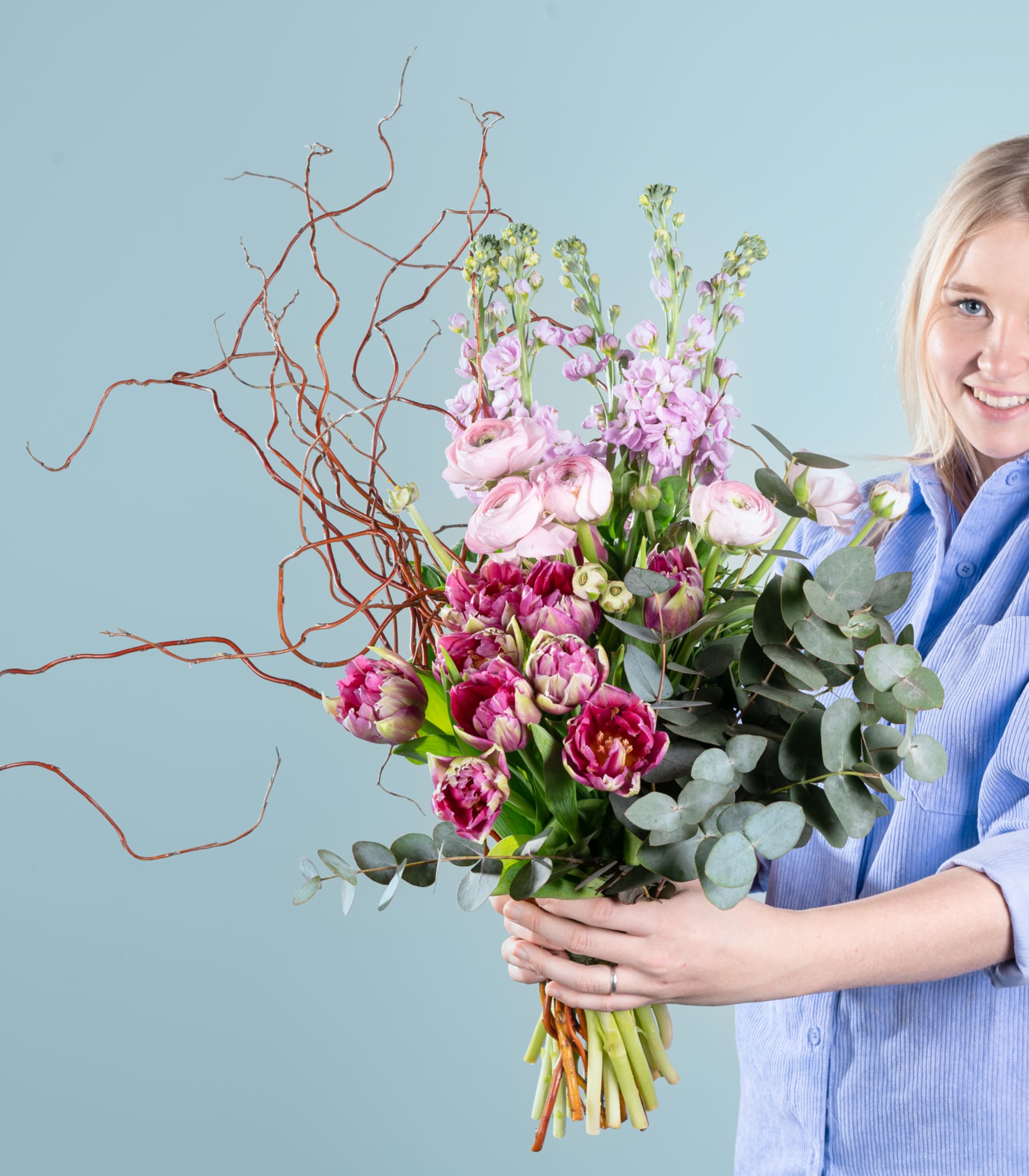 Eine lächelnde Frau in einem blauen Hemd hält einen farbenfrohen Strauß rosa und lila Blumen vor einem schlichten blauen Hintergrund.