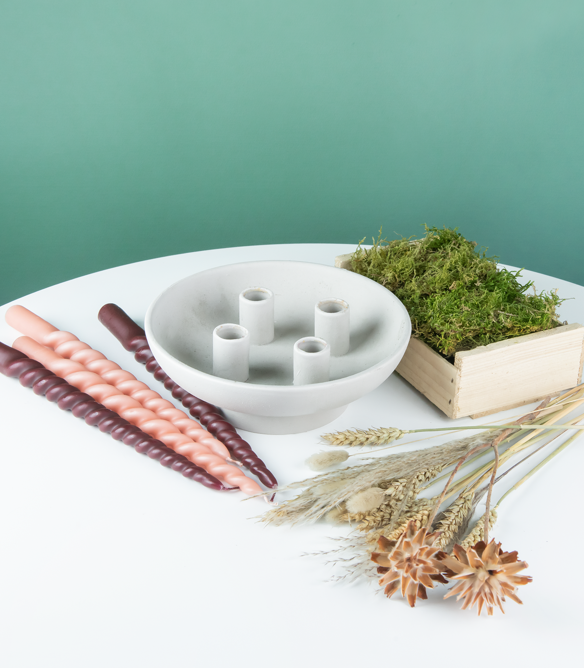 Ein weißer Keramikteller mit verschiedenen Trockenblumen, eine mit Moos gefüllte Holzkiste und ein Satz Holzstäbe auf einer schlichten weißen Fläche vor einem mintgrünen Hintergrund.