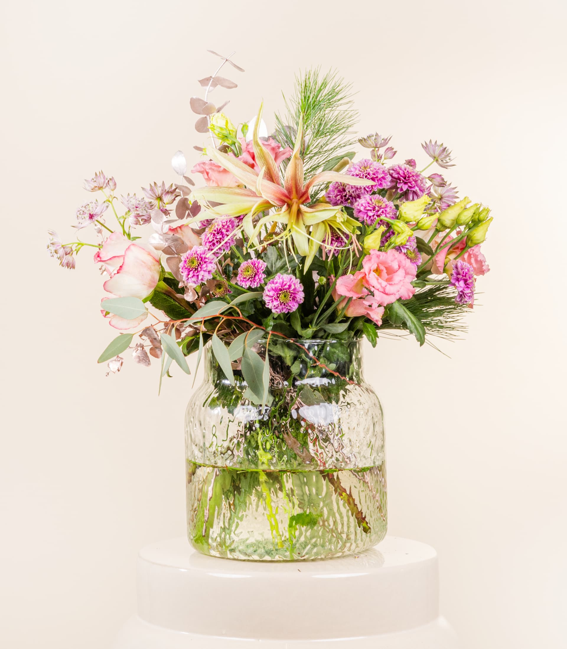 Ein lebendiger Strauß verschiedener bunter Blumen, darunter rosa Rosen, lila Pompons und grünes Laub, wird in einer Glasvase vor einem schlichten, hellen Hintergrund präsentiert. (Beschreibungstext KI-generiert)