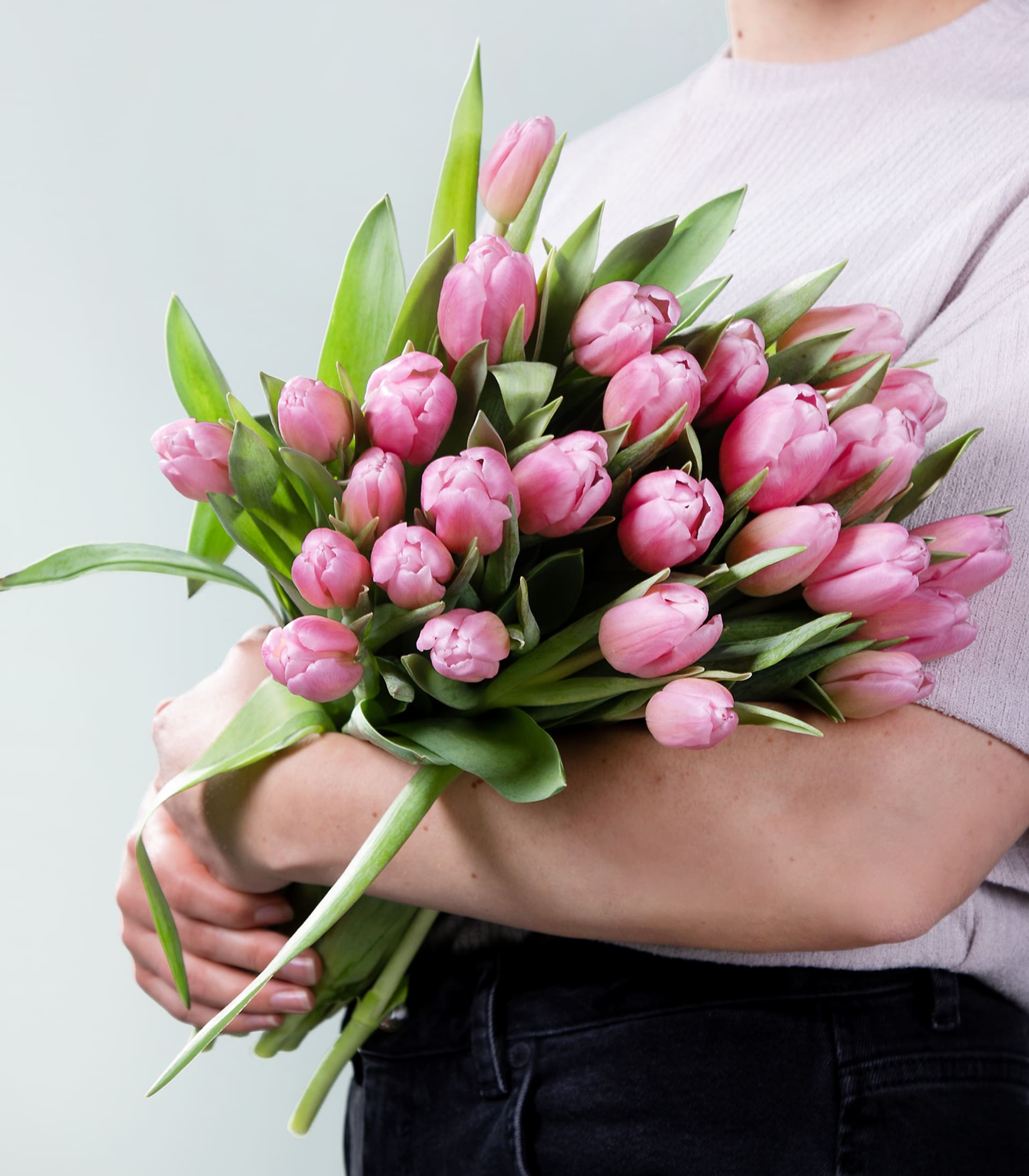 Ein Strauß leuchtend rosa Tulpen wird in den Armen einer Person gehalten, die vor einem schlichten Hintergrund einen hellen Pullover trägt.