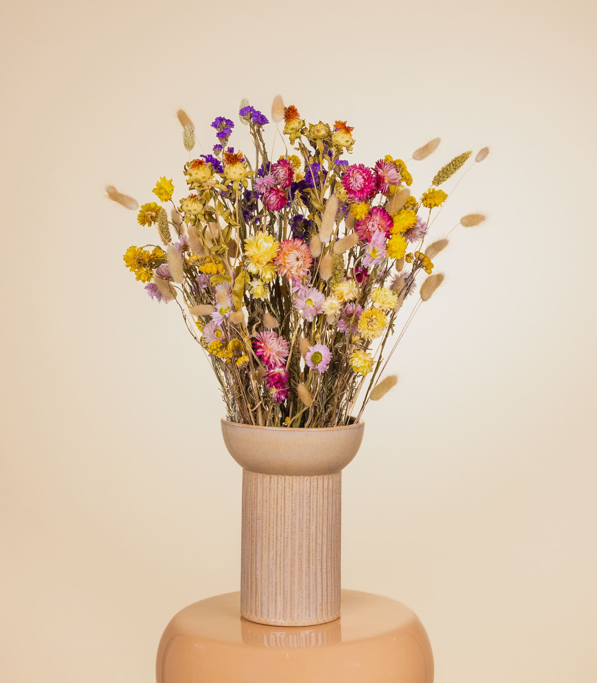 Ein Strauß leuchtender Trockenblumen in verschiedenen Farbtönen, darunter Gelb, Rosa und Lila, wird in einer einfachen Holzvase vor einem neutralen beigen Hintergrund präsentiert.