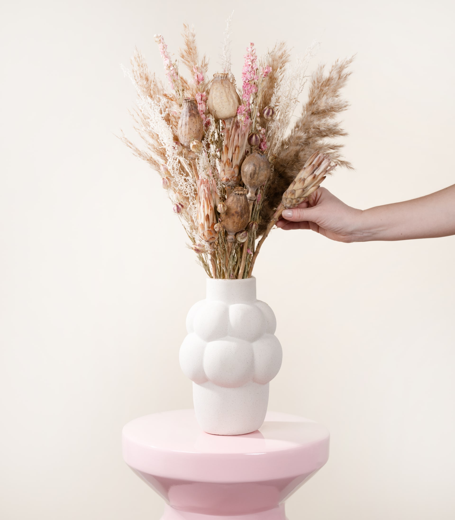 Eine Hand hält einen Strauß getrockneter Blumen in einer weißen Vase vor einem schlichten, hellen Hintergrund.