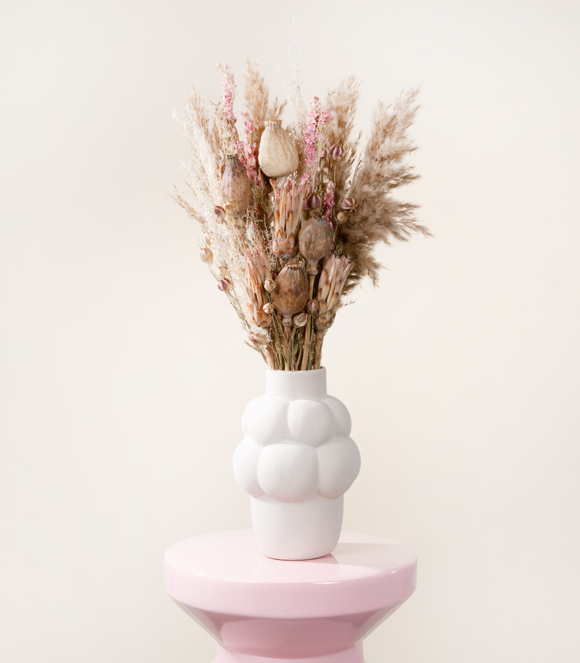 Eine weiße Keramikvase, gefüllt mit einem Strauß getrocknetem Pampasgras und anderen natürlichen Elementen, steht auf einem blassrosa Sockel vor einem schlichten weißen Hintergrund.