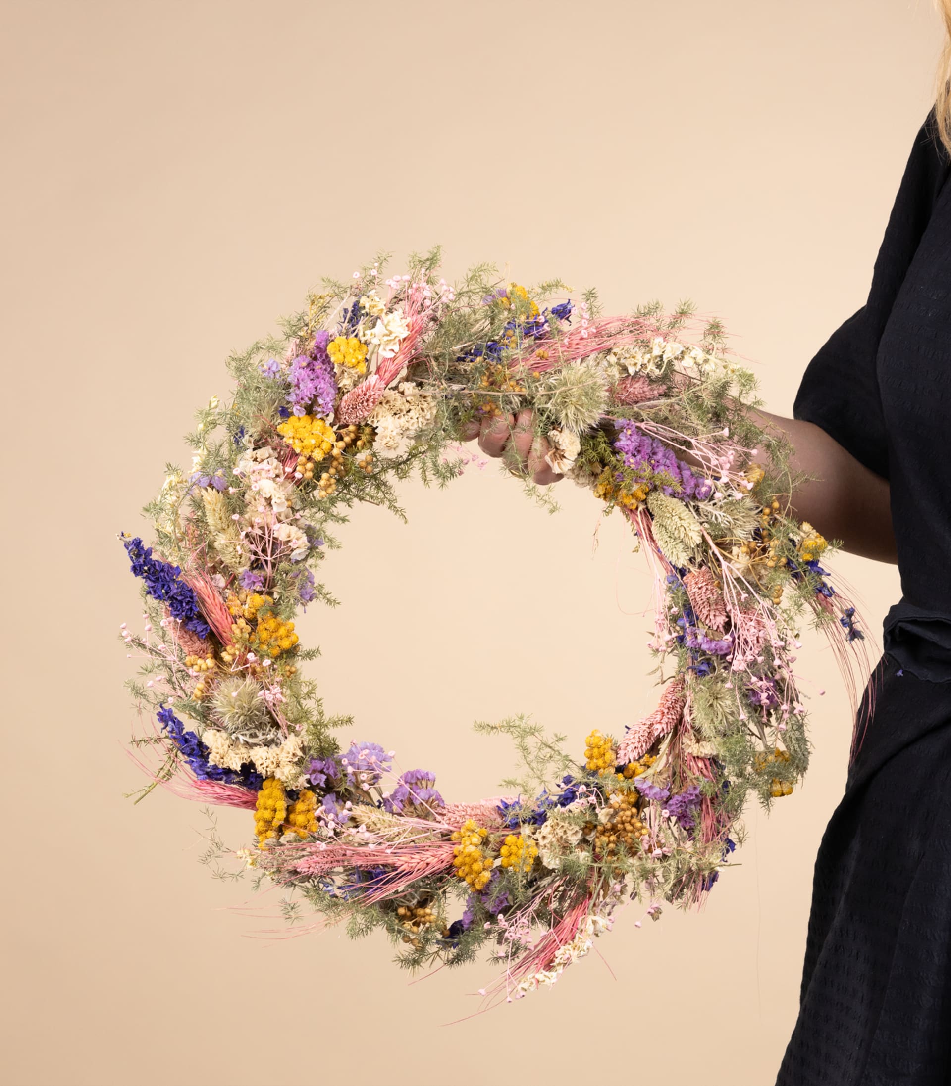 Ein leuchtend bunter Kranz aus verschiedenen Blumen und Blättern wird von einer Person in dunklem Outfit vor einem schlichten, hellen Hintergrund gehalten.