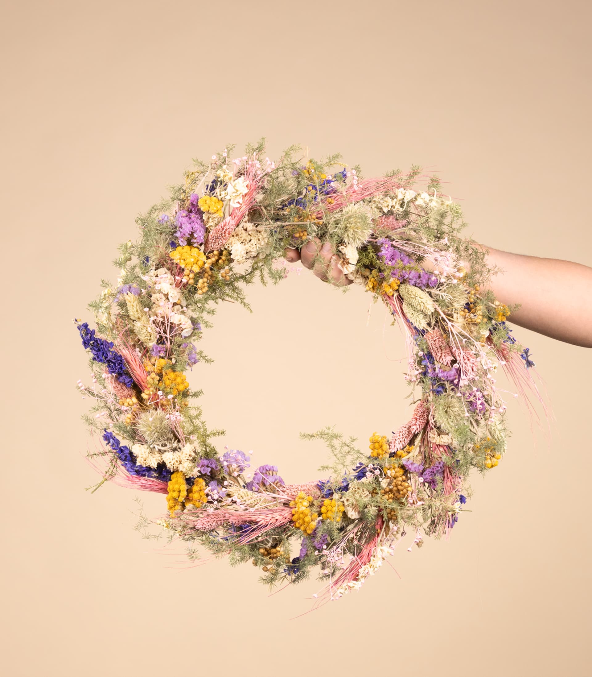 Ein lebendiger, farbenfroher Kranz aus verschiedenen Wildblumen und Grünpflanzen wird von einer Hand vor einem neutralen beigen Hintergrund gehalten.