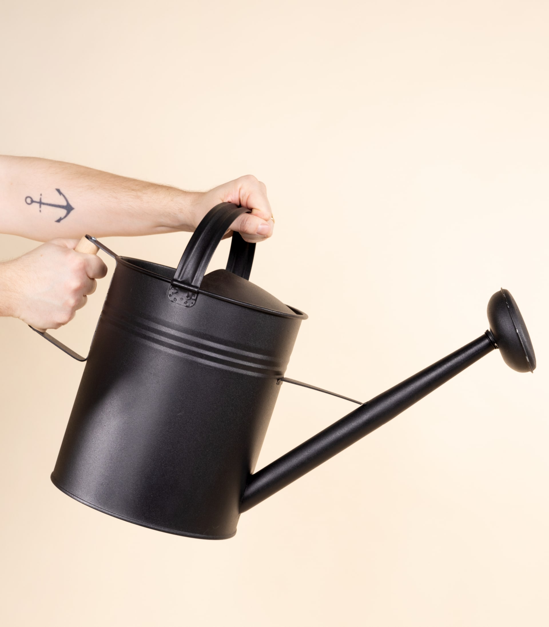 Eine schwarze Gießkanne aus Metall wird von einer Person vor einem schlichten beigen Hintergrund in den Händen gehalten.