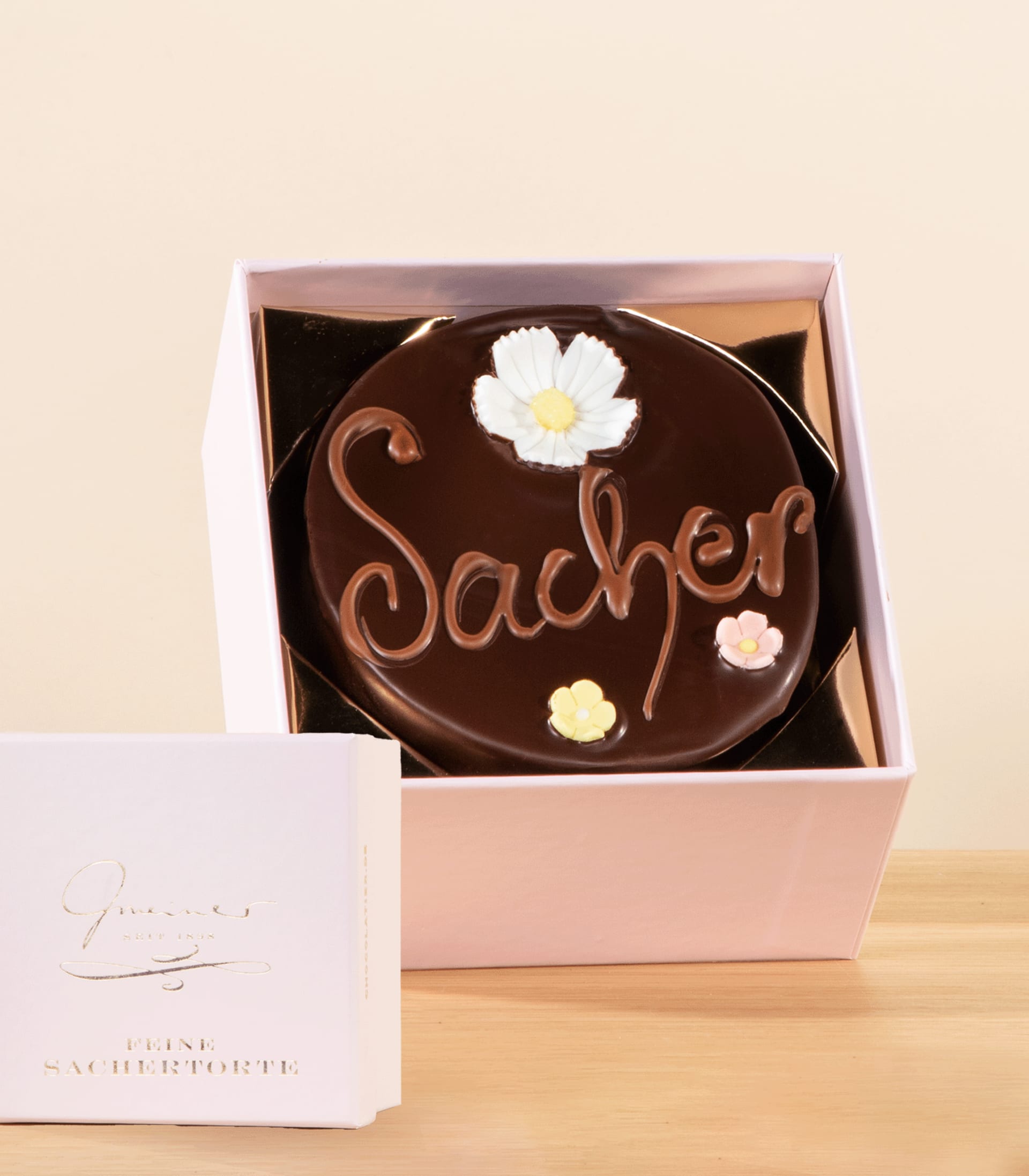 Eine Schokoladentorte mit der Aufschrift „Sacher“, dekoriert mit weißen Blumen, steht in einer rosa Schachtel auf einer Holzoberfläche.
