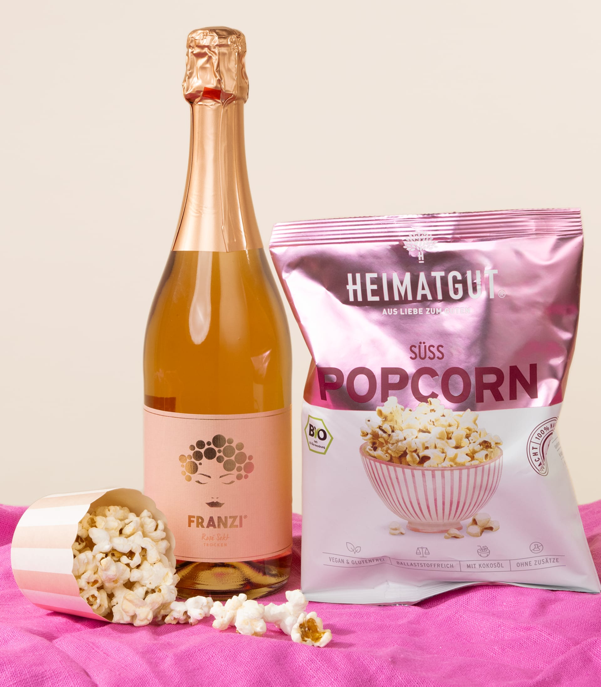 Auf einem rosa Stoffhintergrund sind eine Flasche Sekt, eine Tüte süßes Popcorn und verstreute Popcornkörner arrangiert.