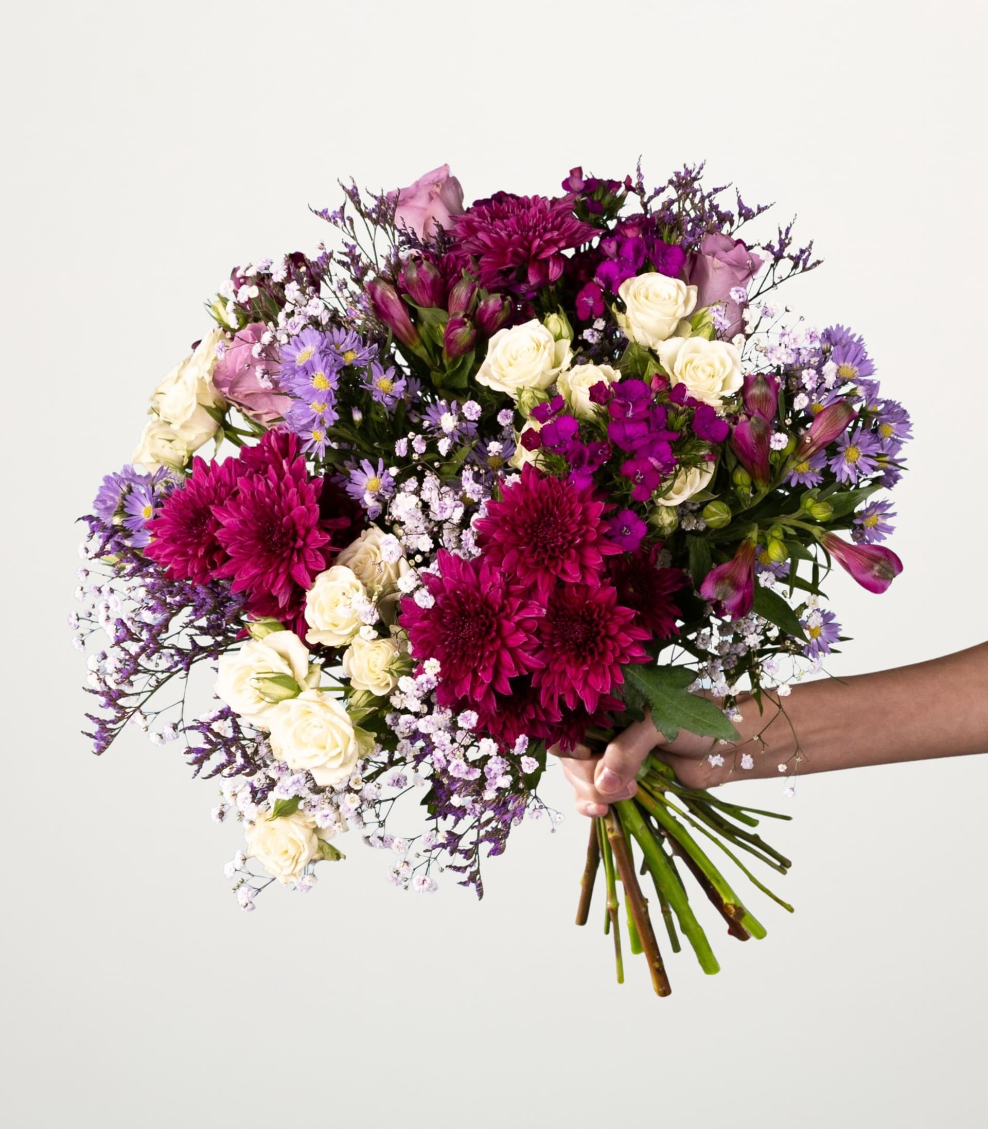 Ein lebendiger Strauß verschiedener bunter Blumen, darunter rosa und violette Chrysanthemen, weiße Rosen und zarte violette und weiße Blumen, die eine Person vor einem schlichten weißen Hintergrund in den Händen hält.
