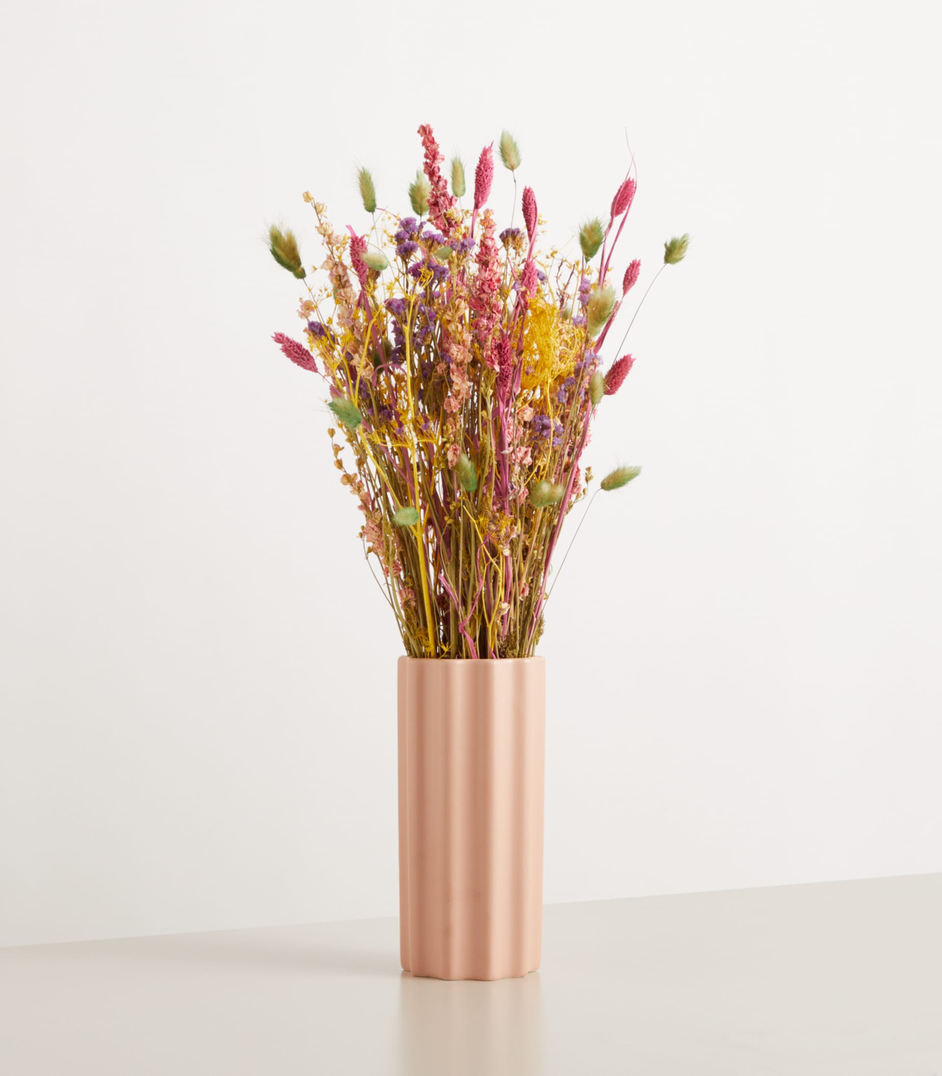 Ein Strauß leuchtender Trockenblumen in einer metallisch rosa Vase steht vor einem schlichten weißen Hintergrund.