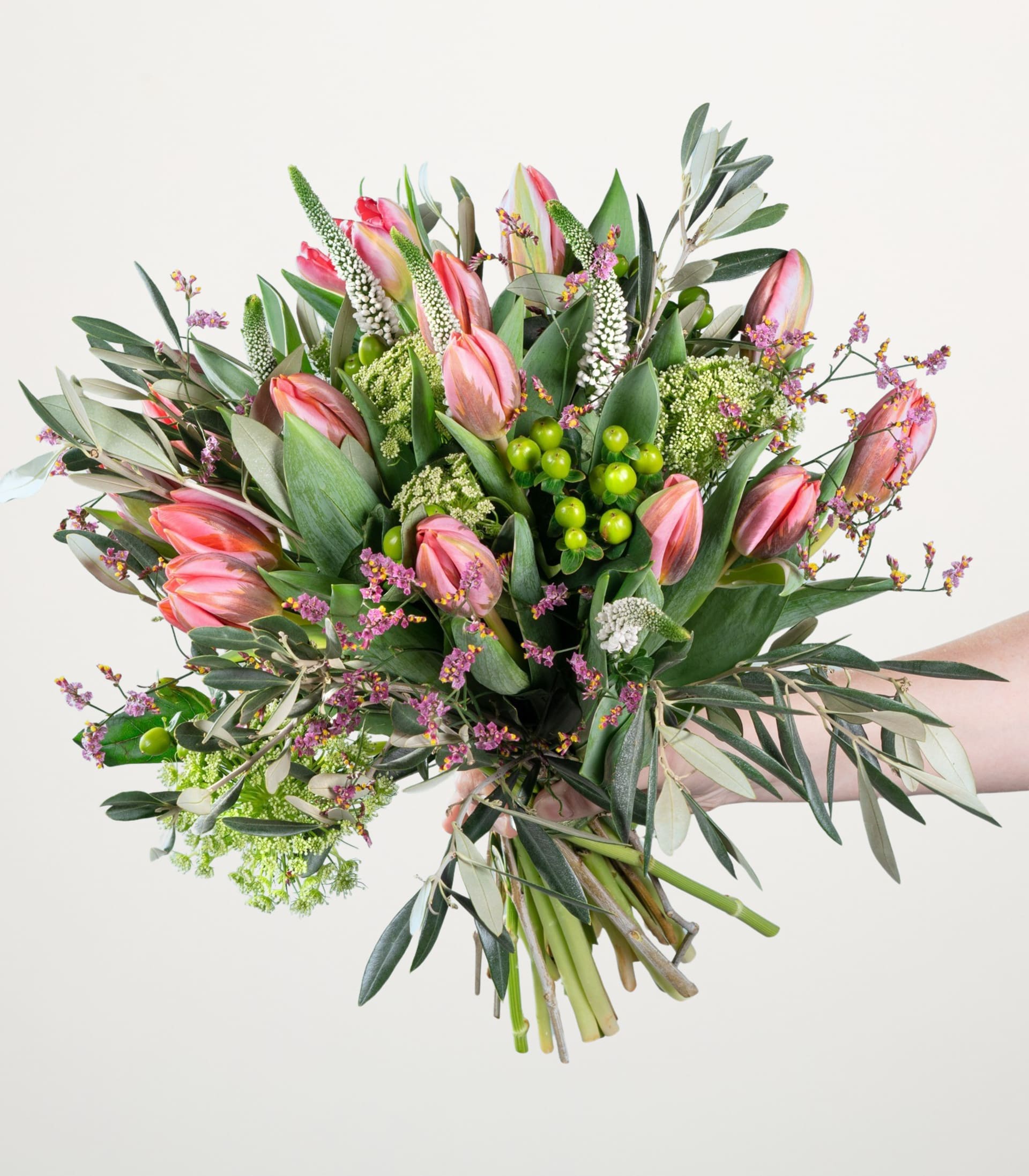 Ein leuchtender Strauß aus rosa Tulpen, grünem Laub und anderen bunten Blumen, arrangiert in einem Handdisplay vor einem schlichten weißen Hintergrund.