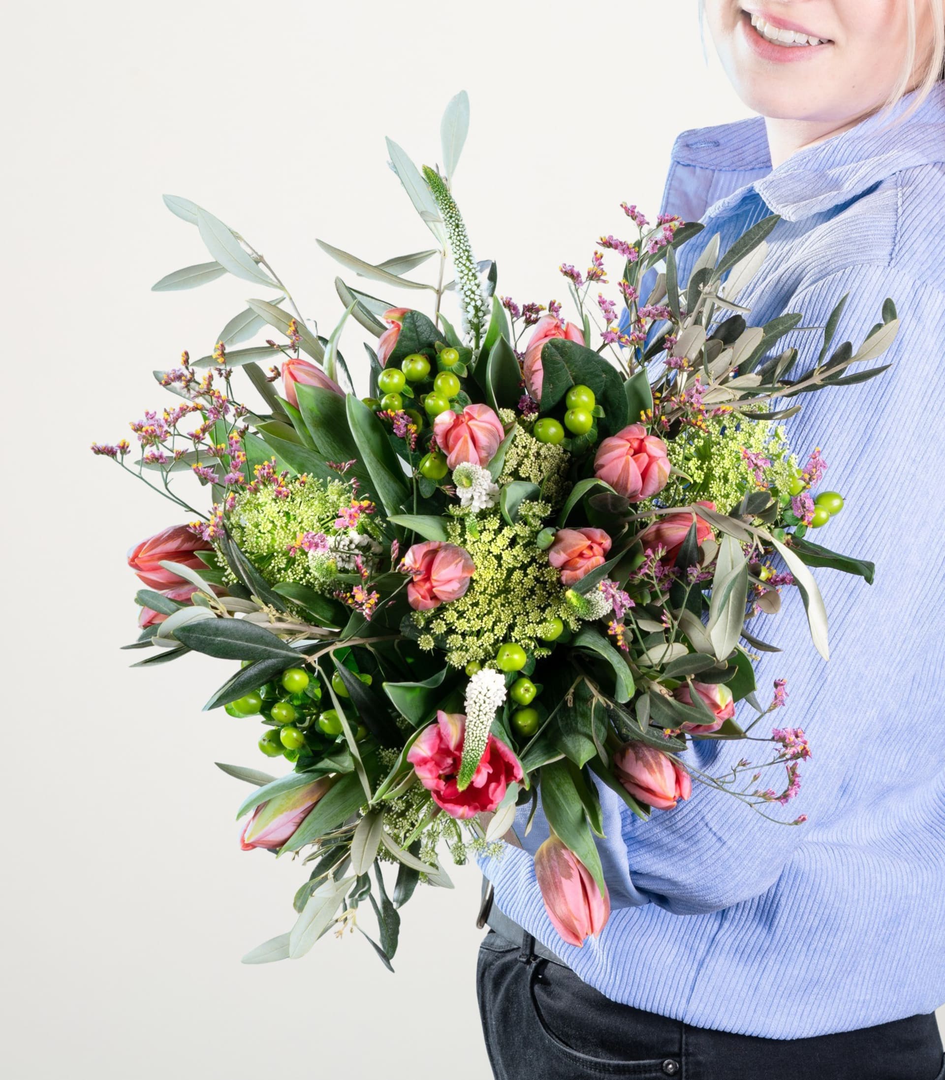 Ein leuchtender Strauß bunter Blumen, darunter rosa Rosen und grünes Laub, wird von einer Person in einem blauen Pullover vor einem schlichten weißen Hintergrund gehalten.