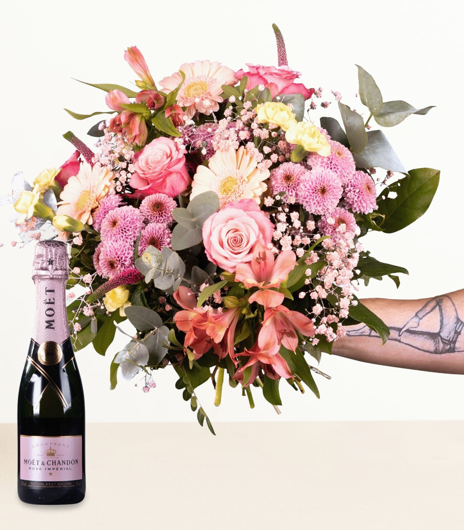 Ein farbenfroher Strauß rosa, gelber und weißer Blumen steht neben einer Flasche Sekt vor einem schlichten weißen Hintergrund.